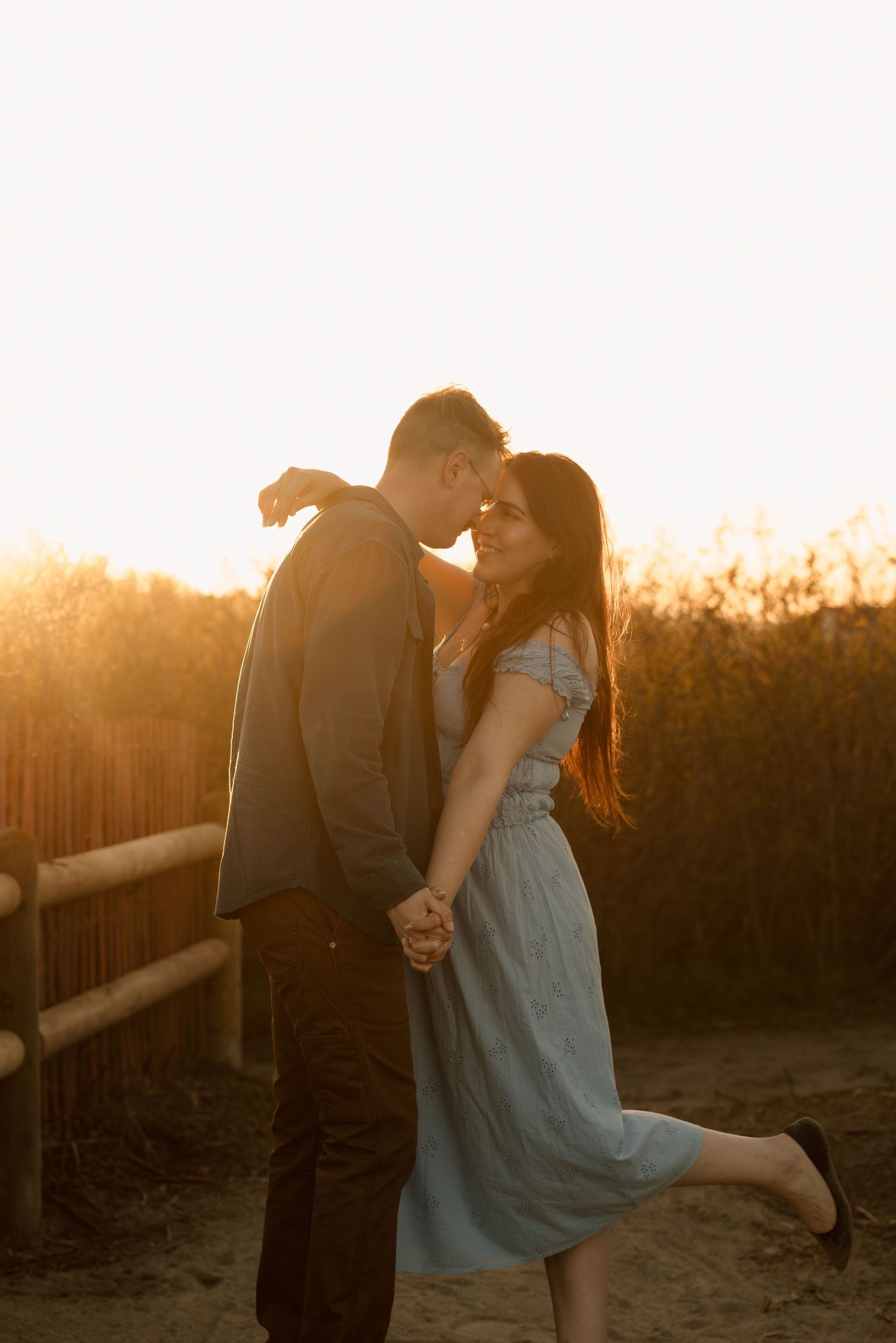Couple sharing a romantic moment at golden hour sunset, woman in light blue dress lifting her leg.