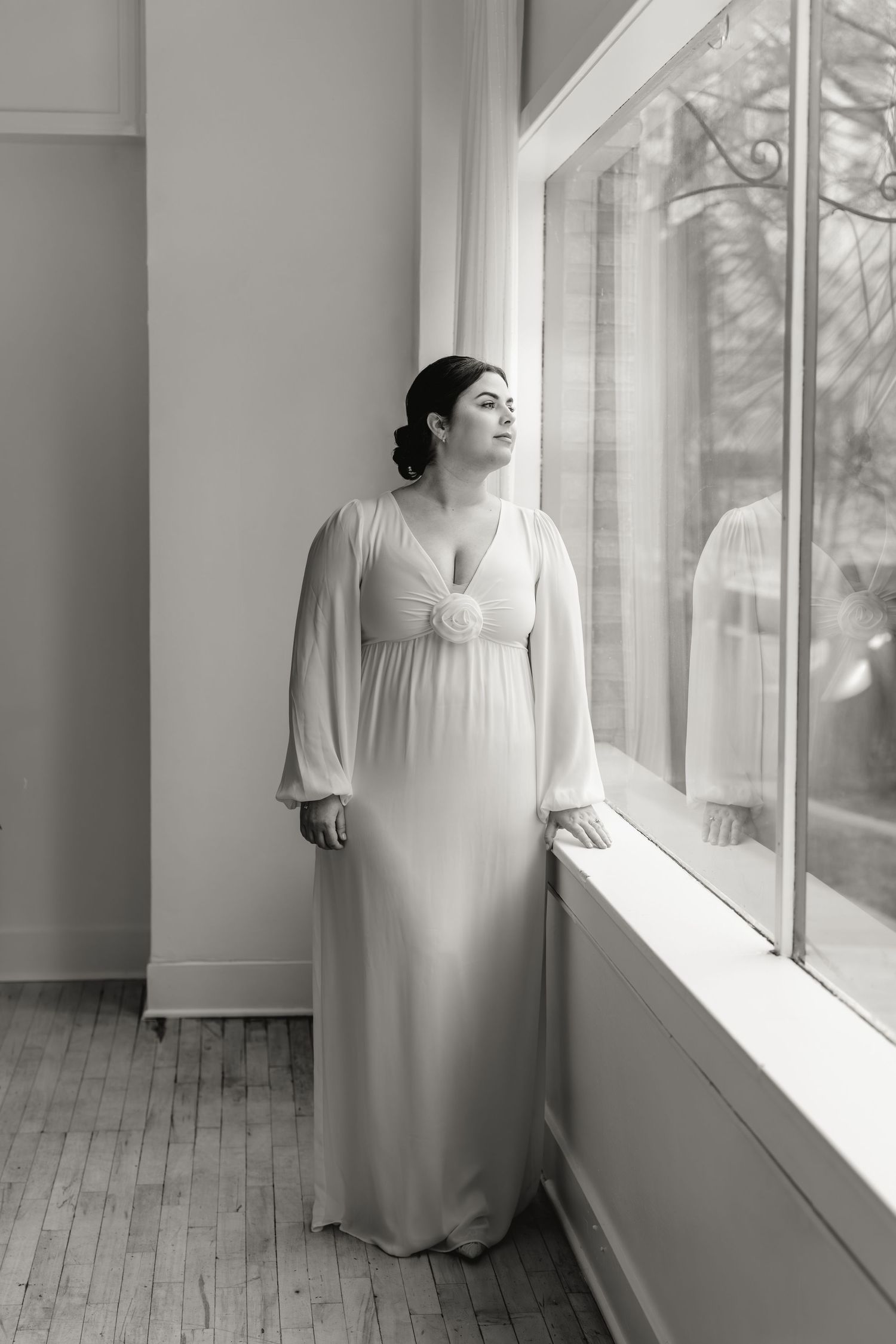 A person in a flowing white dress stands by large windows in a bright minimalist interior space.