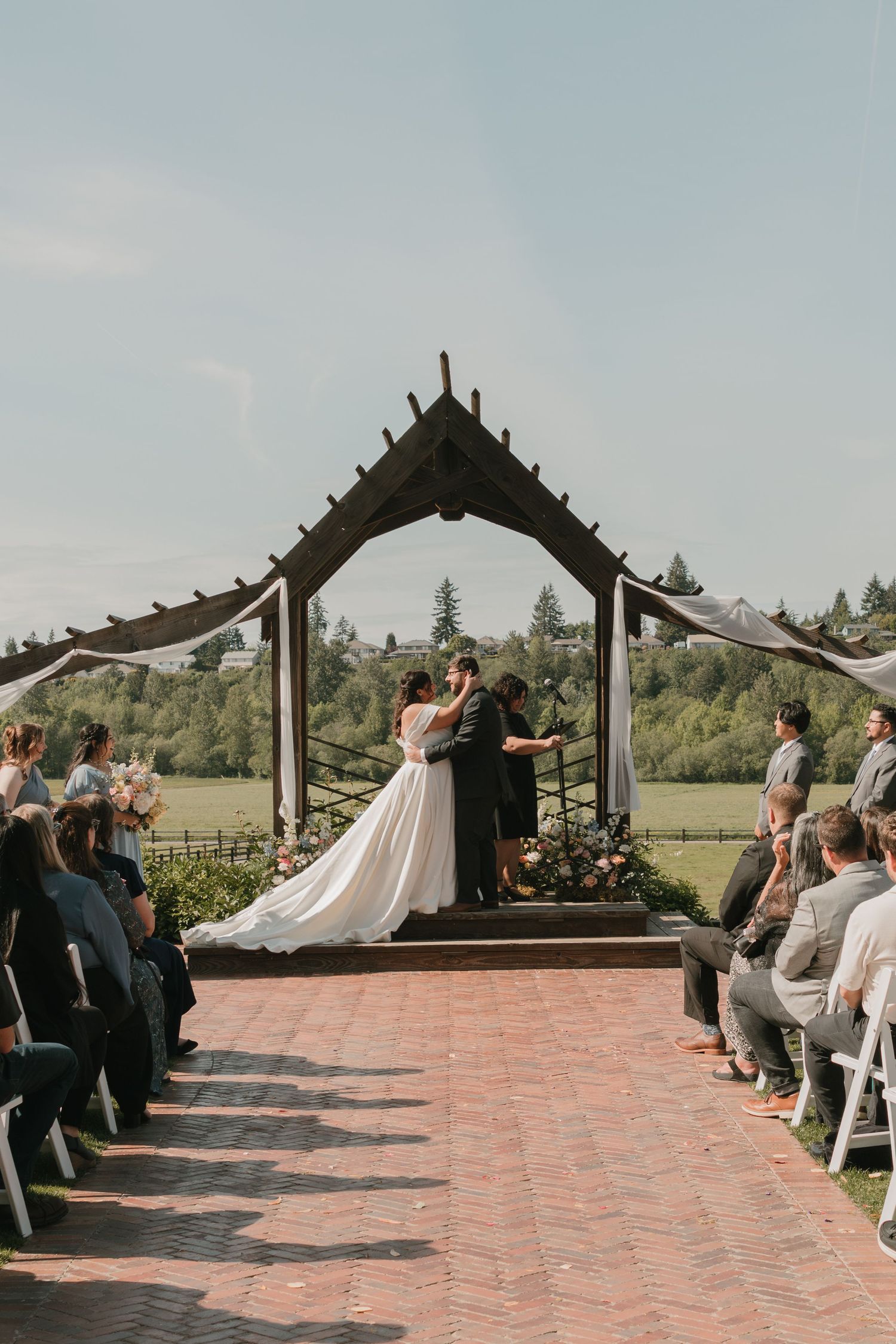 An outdoor wedding ceremony takes place under a rustic wooden arch on a brick pathway surrounded by guests and greenery.