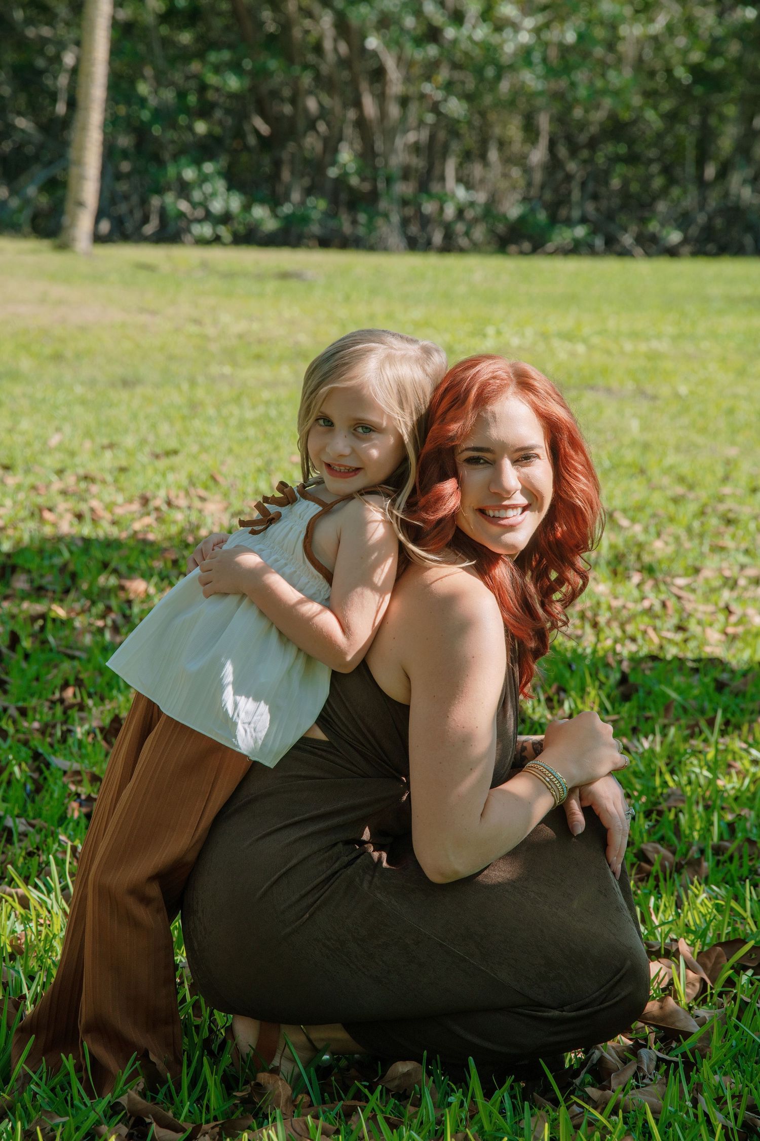 A redhead woman and young blonde girl hug and smile together on green grass in a sunny park.