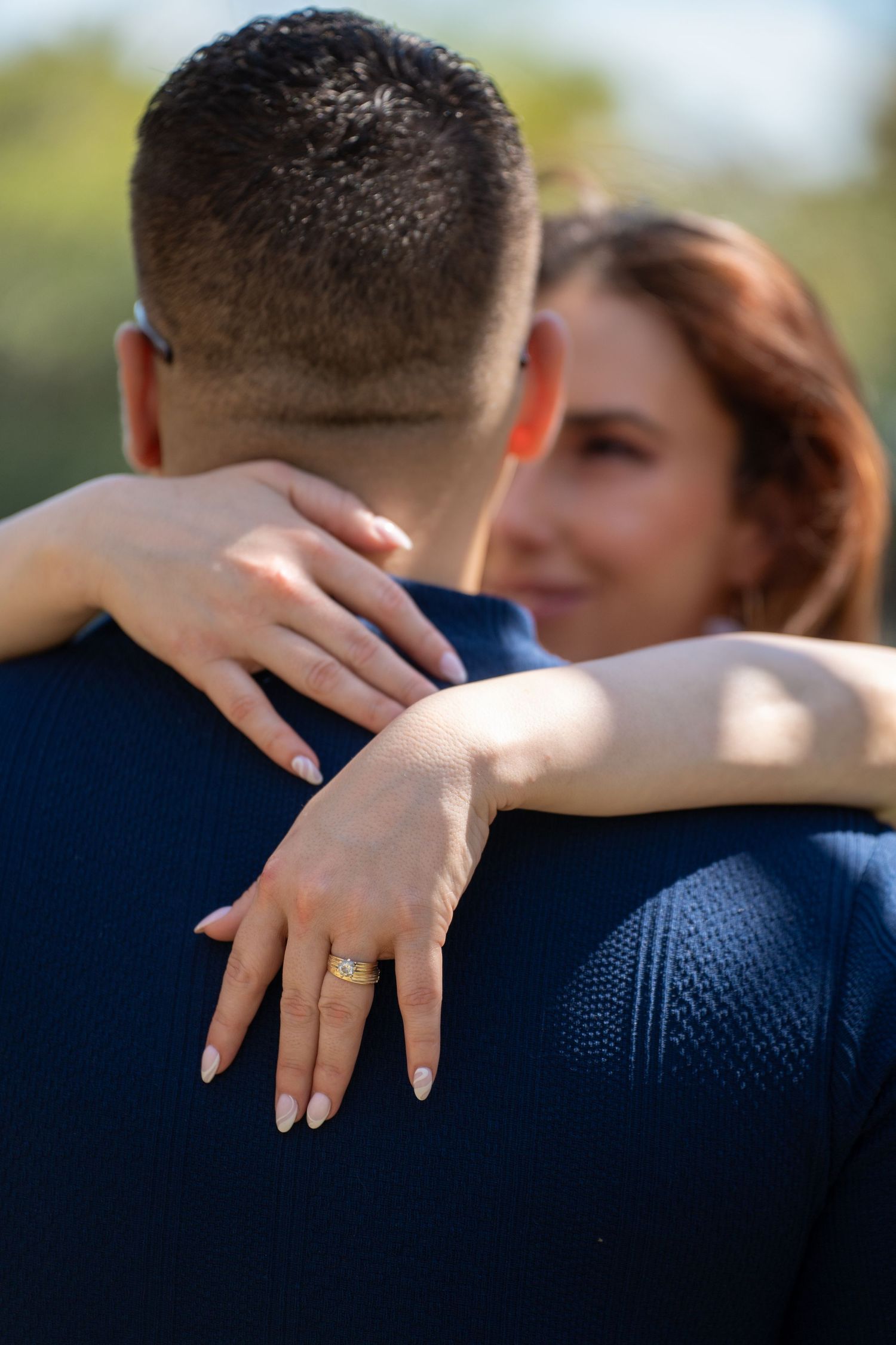 Couple embracing outdoors, woman wearing gold engagement ring, man in navy blue sweater, romantic moment captured from behind.