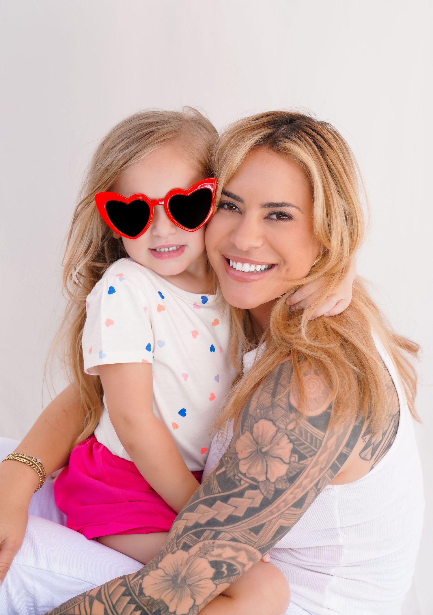 Smiling blonde woman with tattoos hugs young girl wearing red heart-shaped sunglasses and pink skirt against white background.