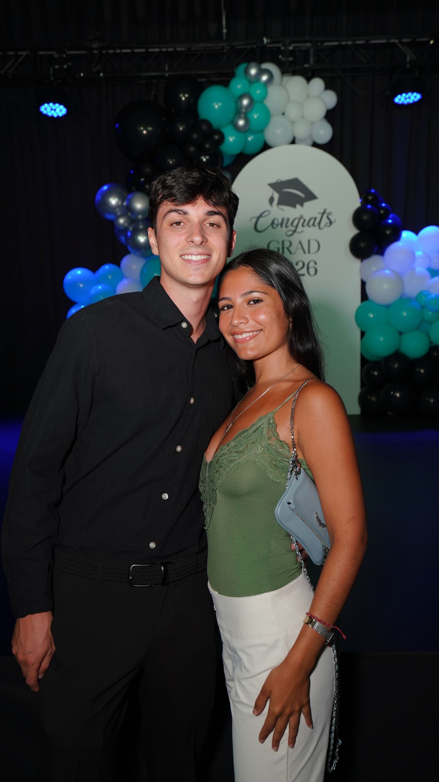 Two young adults pose together at a Congrats Grad 2026 celebration with teal and black balloons.