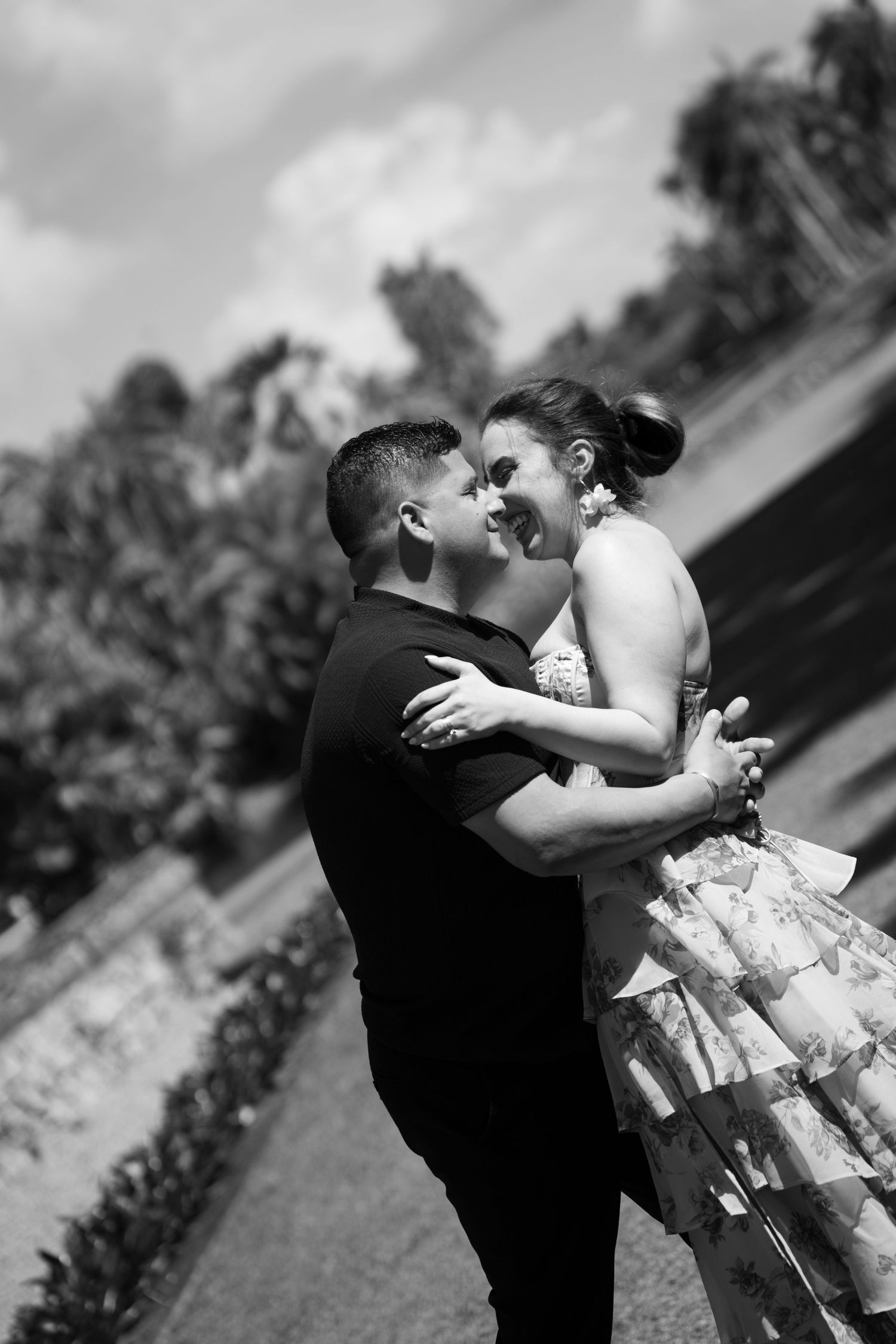 Black and white photo of a romantic couple embracing and smiling at each other outdoors near a garden path.