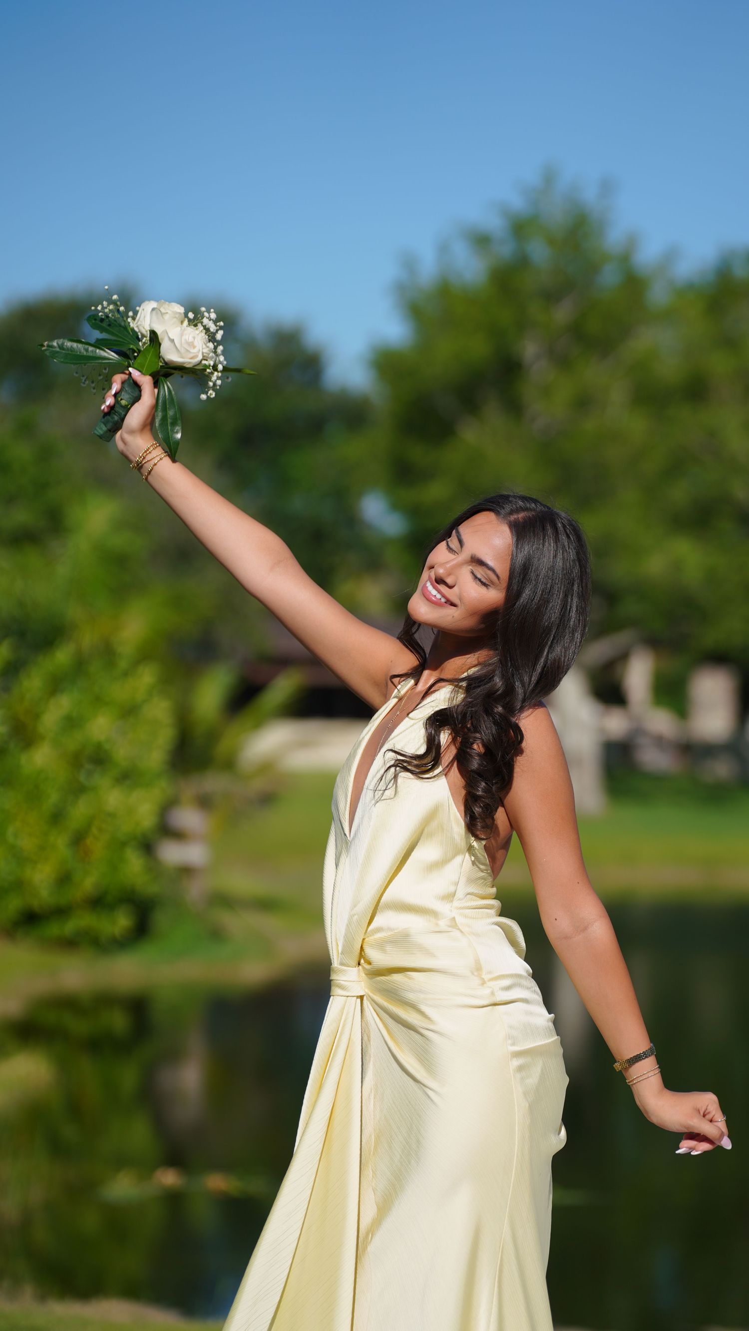 Smiling woman in yellow satin dress holding white flower bouquet outdoors near pond with green trees.