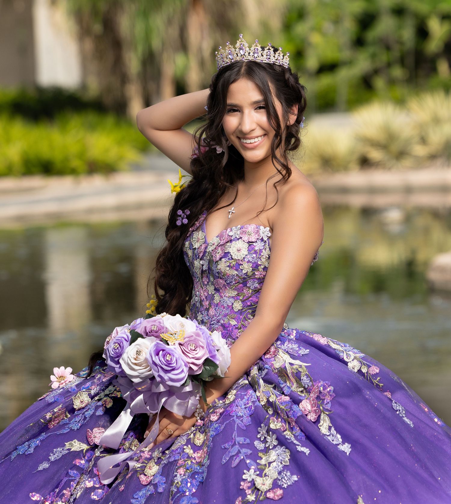 Young woman in purple floral quinceañera ball gown with tiara holding bouquet, posing outdoors near fountain.