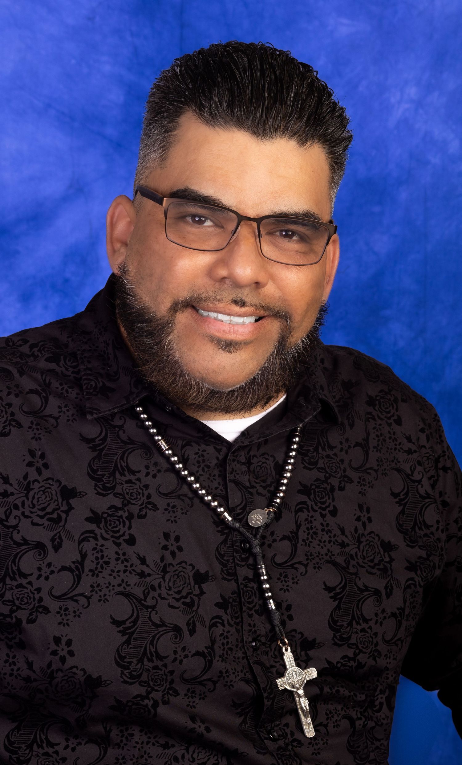 Man with glasses and beard wearing black floral shirt and cross necklace poses for professional portrait photo.