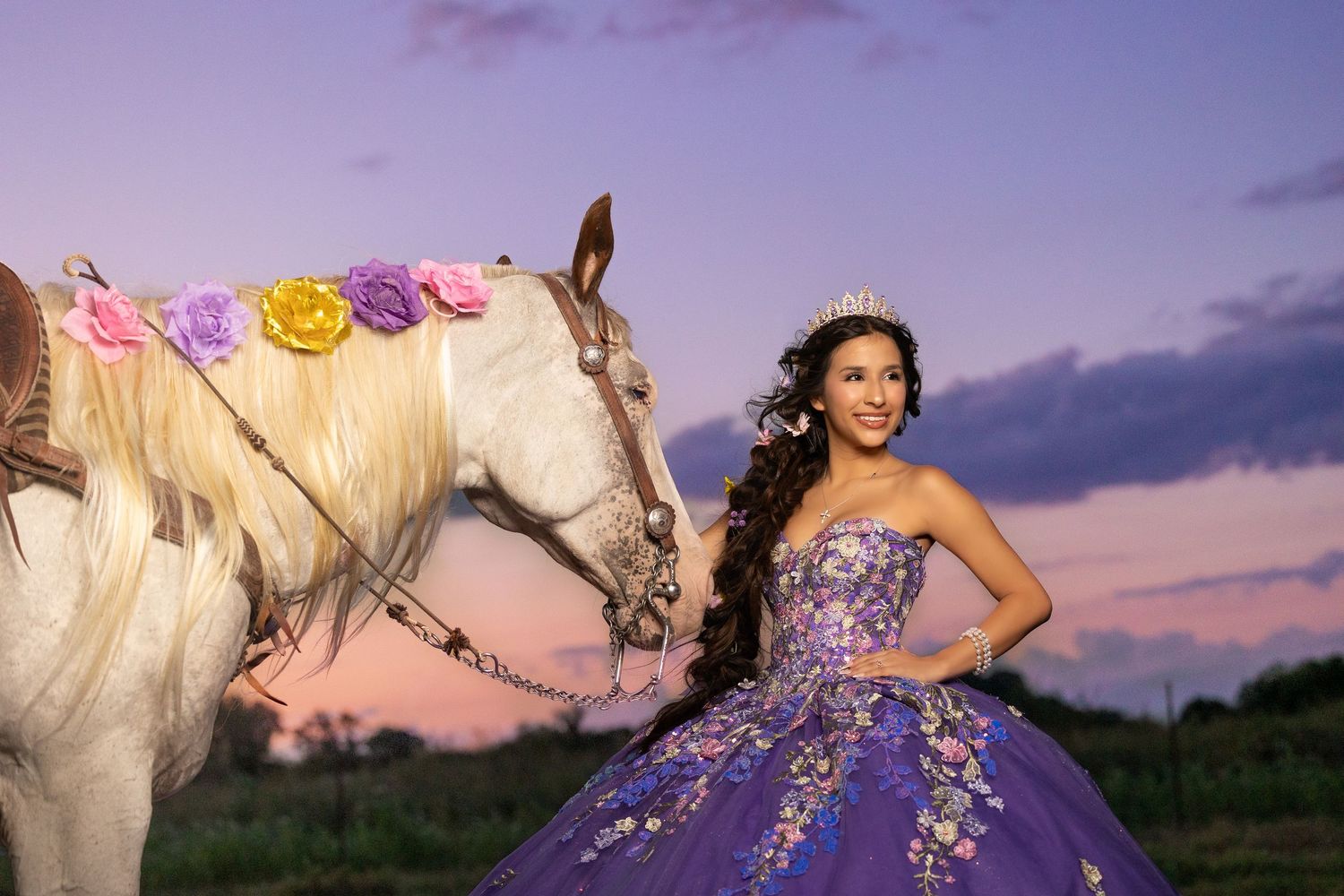 Young woman in purple floral quinceañera gown and tiara poses with a white horse adorned with colorful flowers at sunset.