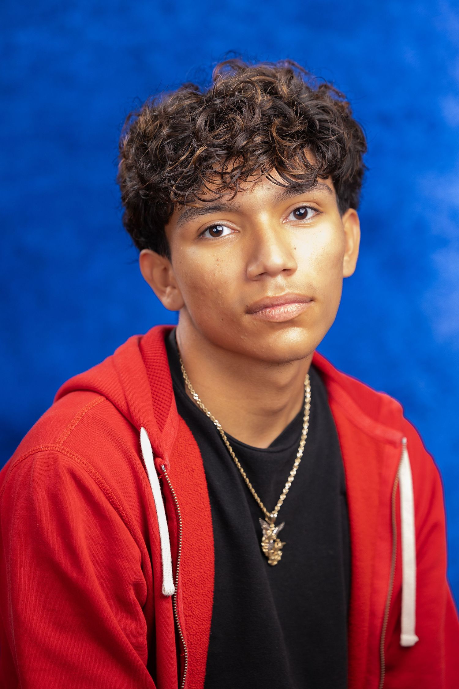Young man with curly hair wearing a red hoodie and gold necklace poses against a blue background.