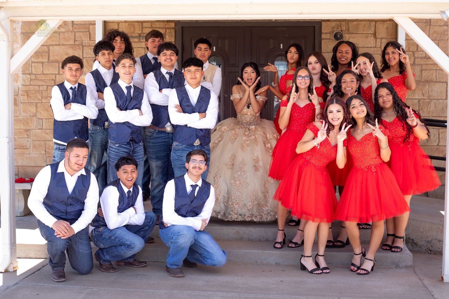 Group of teens in coordinated outfits, boys in navy vests and girls in red dresses, posing together at a quinceañera.