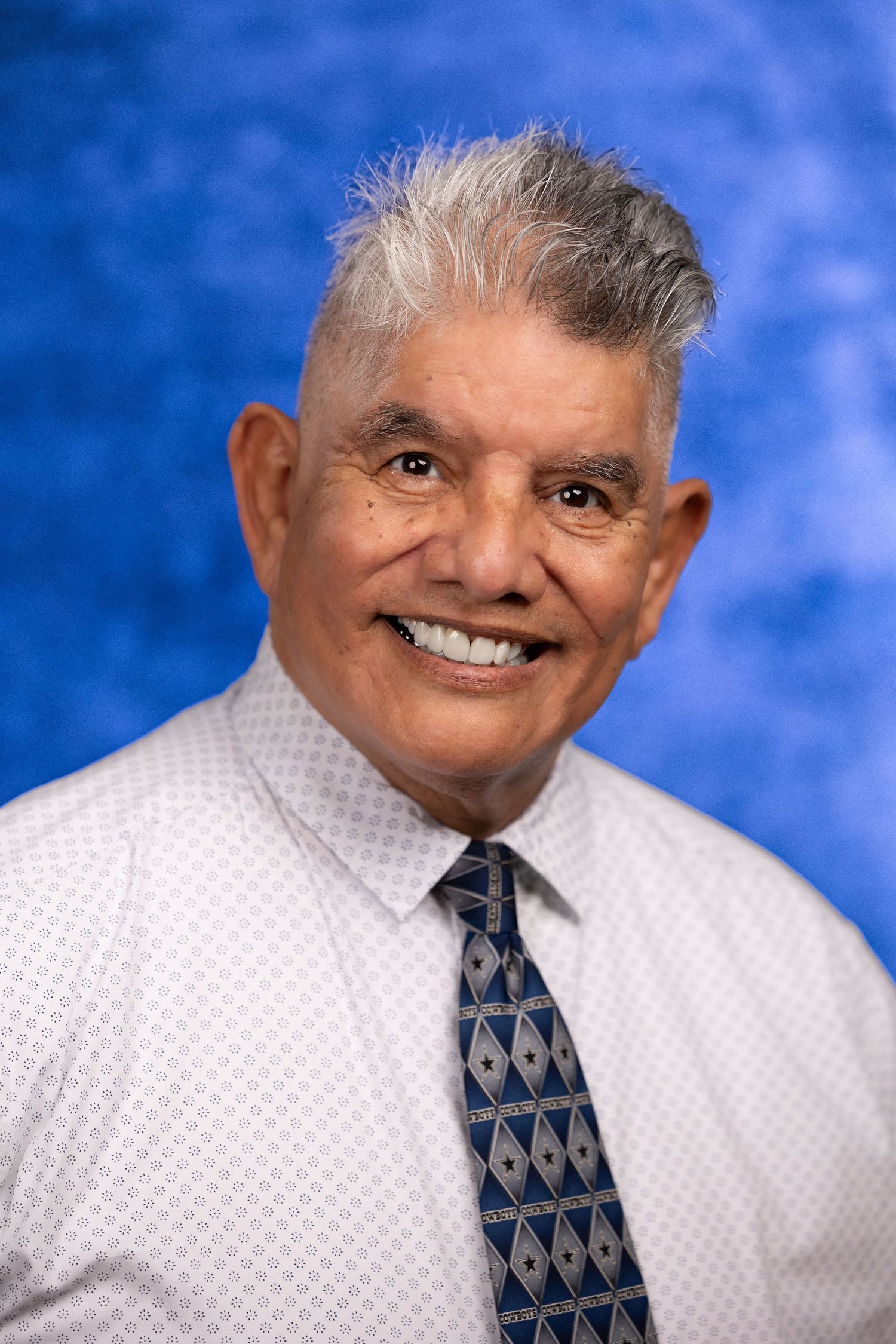 Smiling elderly man with gray hair wearing a white dress shirt and blue diamond-patterned tie against a blue background.