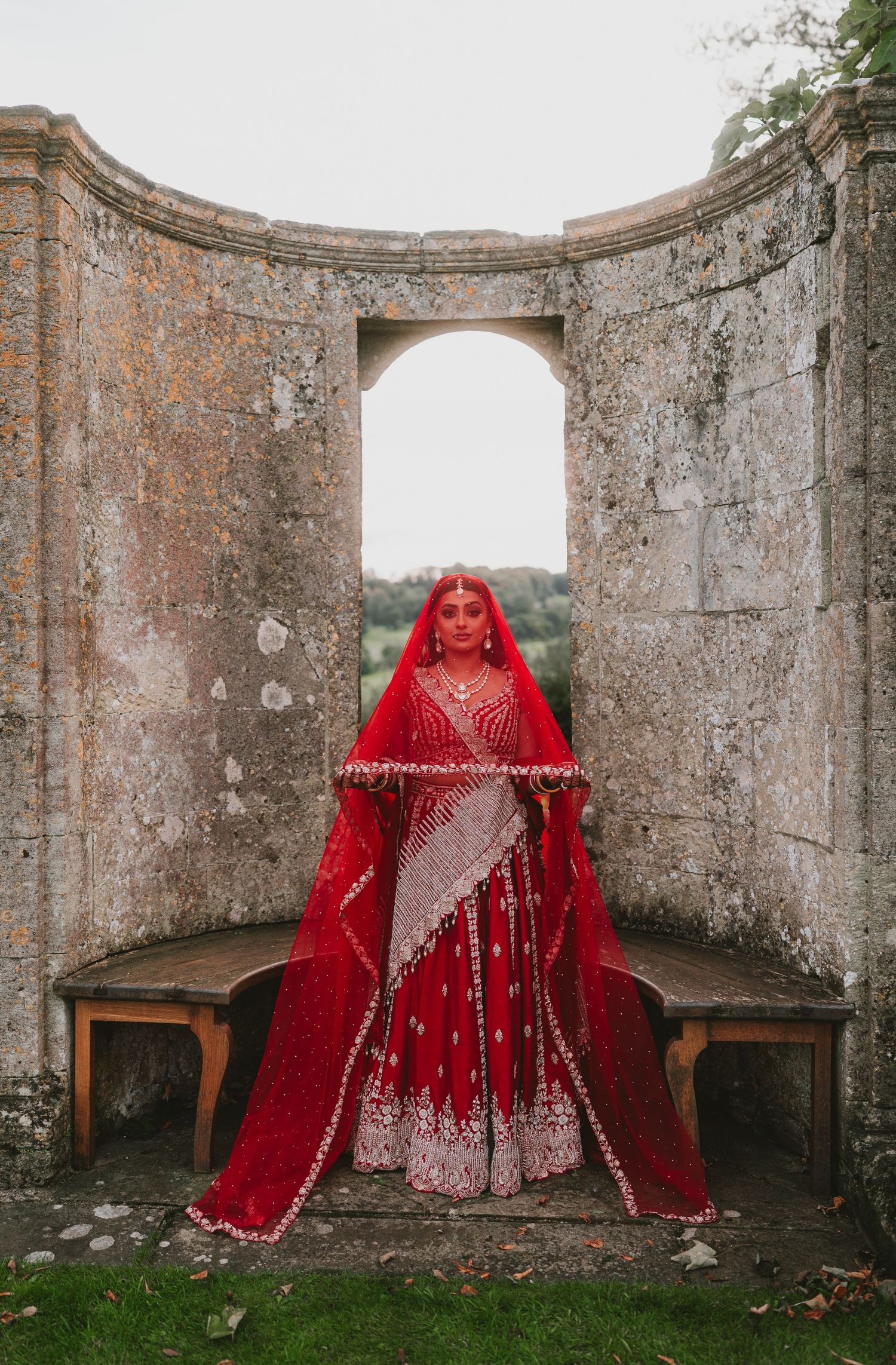 A stunning red and silver traditional bridal lehenga photographed in a stone archway setting.