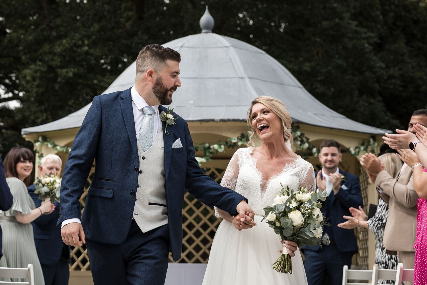 Happy newlyweds walk hand in hand under a white gazebo surrounded by guests during their outdoor wedding ceremony.