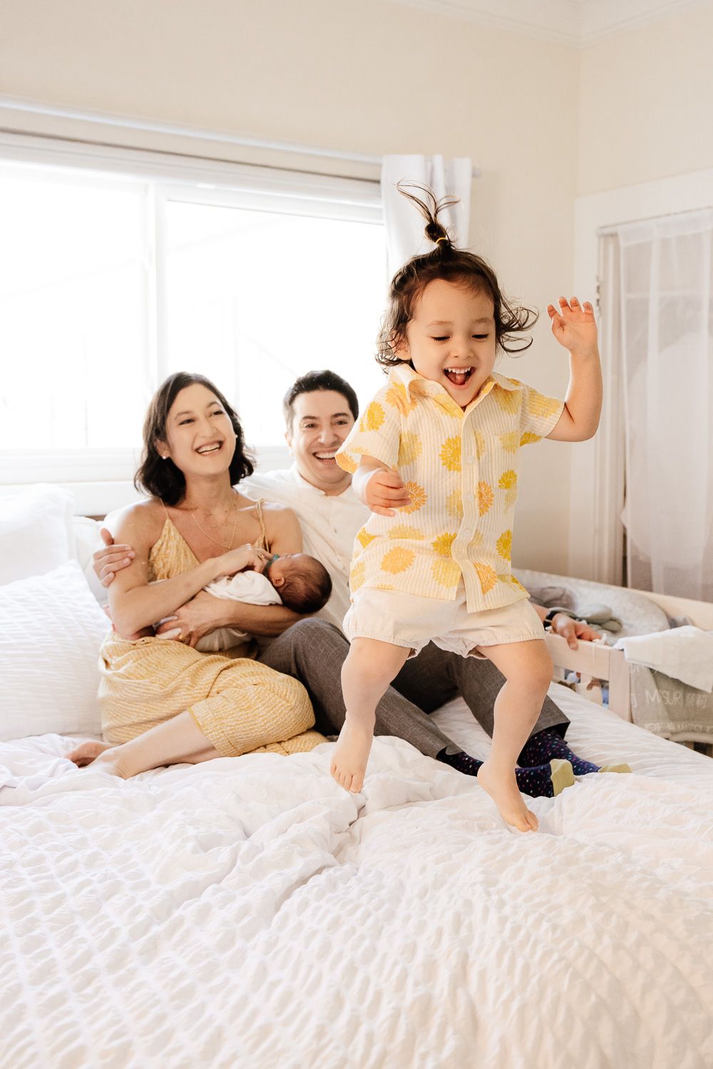 Parents hold their baby at their in-home Bay Area newborn photography session, while their exuberant toddler happily jumps on their bed