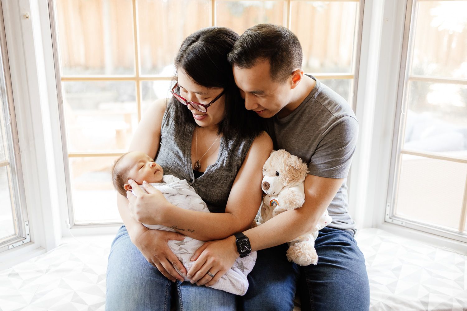 A couple sits in a window nook with beautiful natural light while holding their newborn baby during their in-home newborn photoshoot