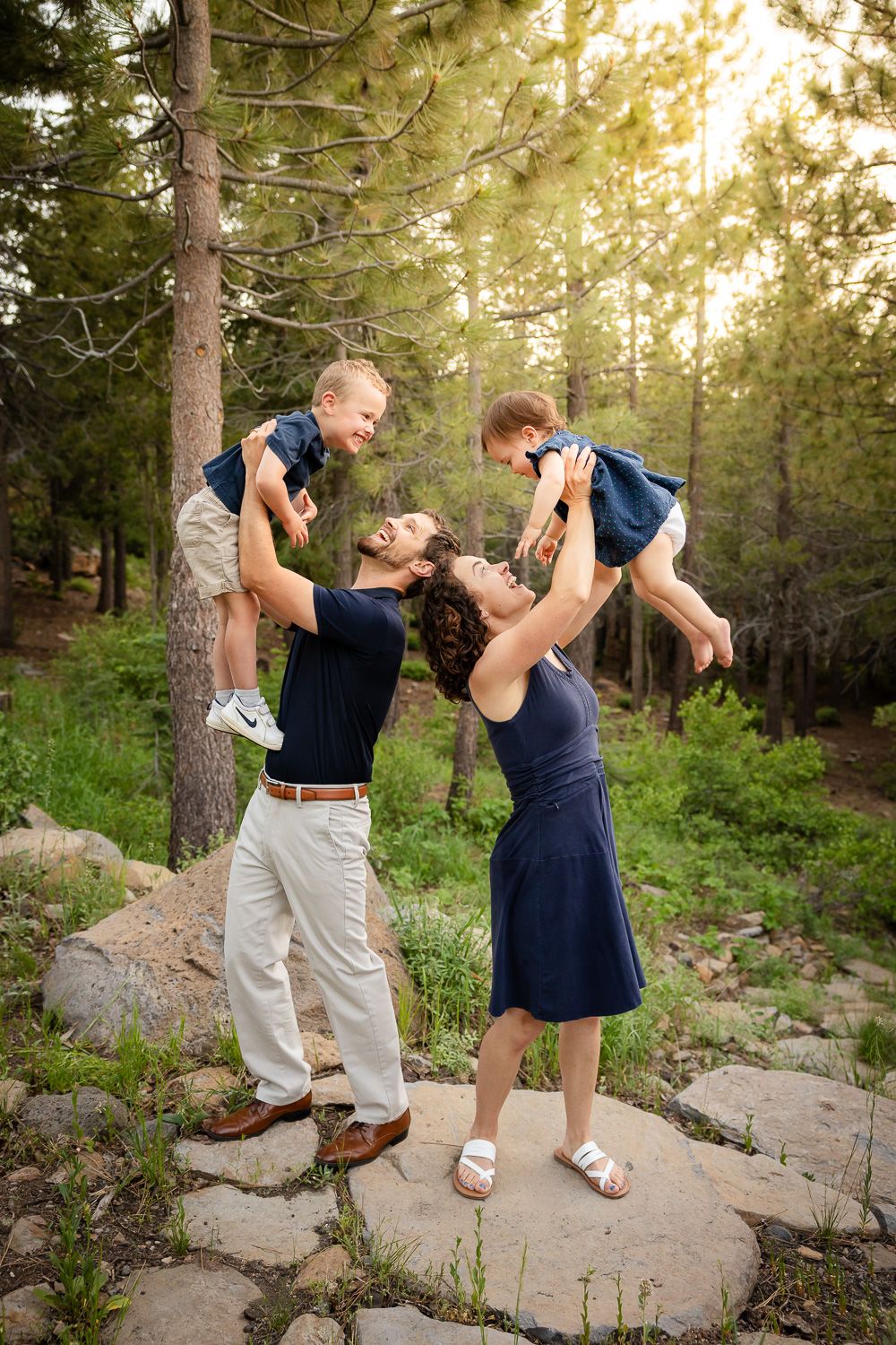 During a family photoshoot outdoors, a mom and dad lift their children skyward playing airplane, much to the children's delight