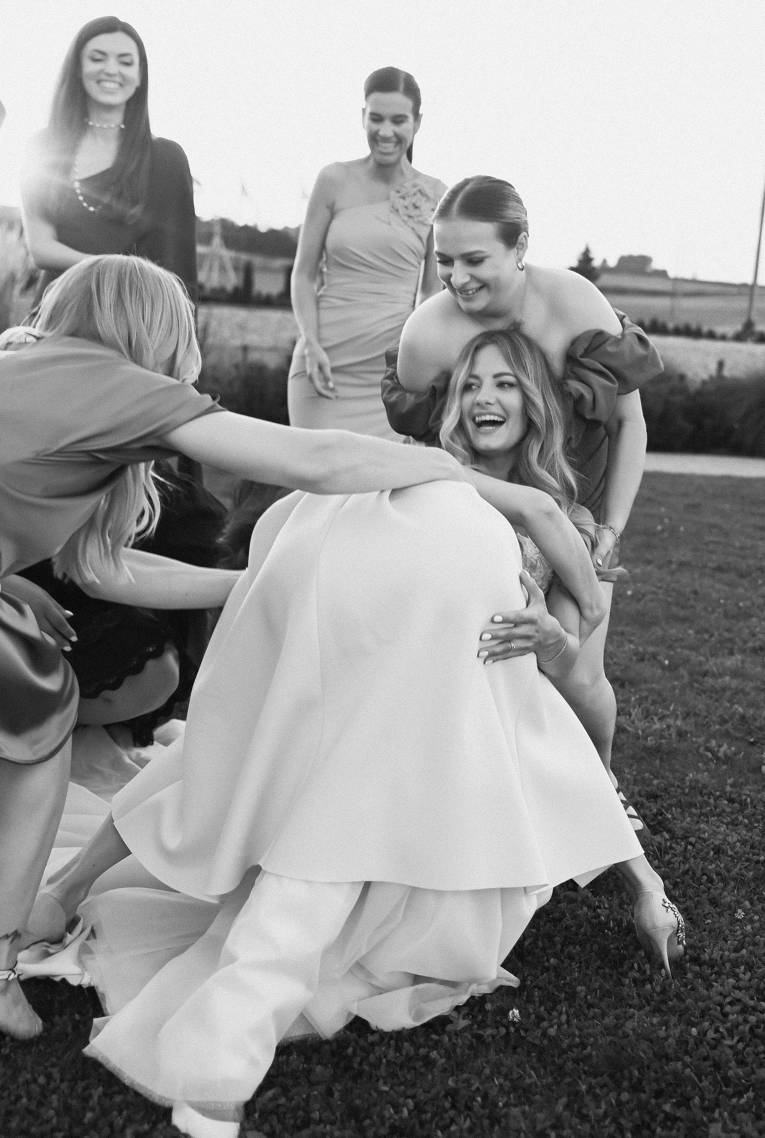 A candid black and white photo showing joyful laughter and playful interactions at an outdoor wedding celebration.