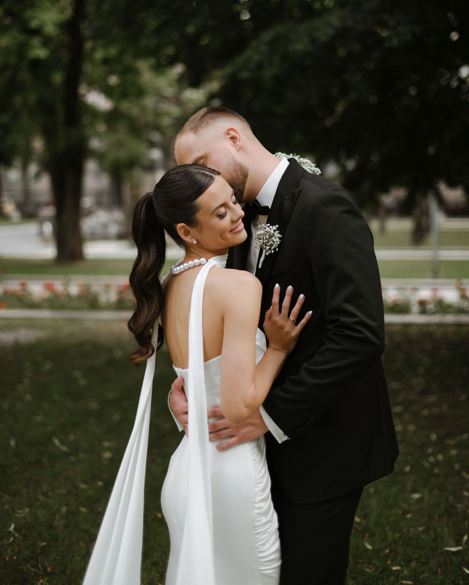 A tender moment between newlyweds embracing outdoors in a garden setting, the bride wearing a flowing white dress with cape detail.