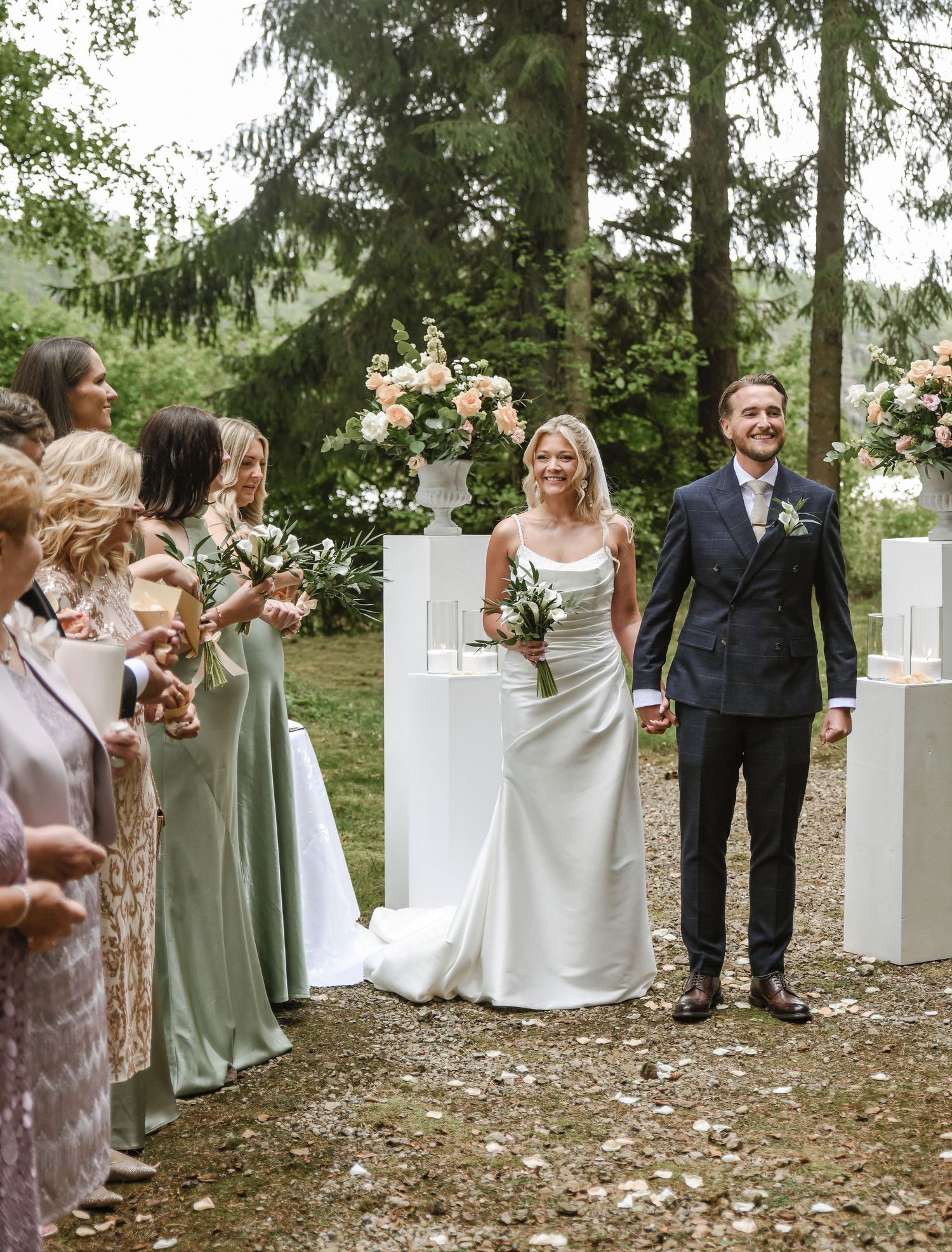 Outdoor forest wedding ceremony with elegant white pedestals and floral arrangements surrounded by bridesmaids in sage green dresses.