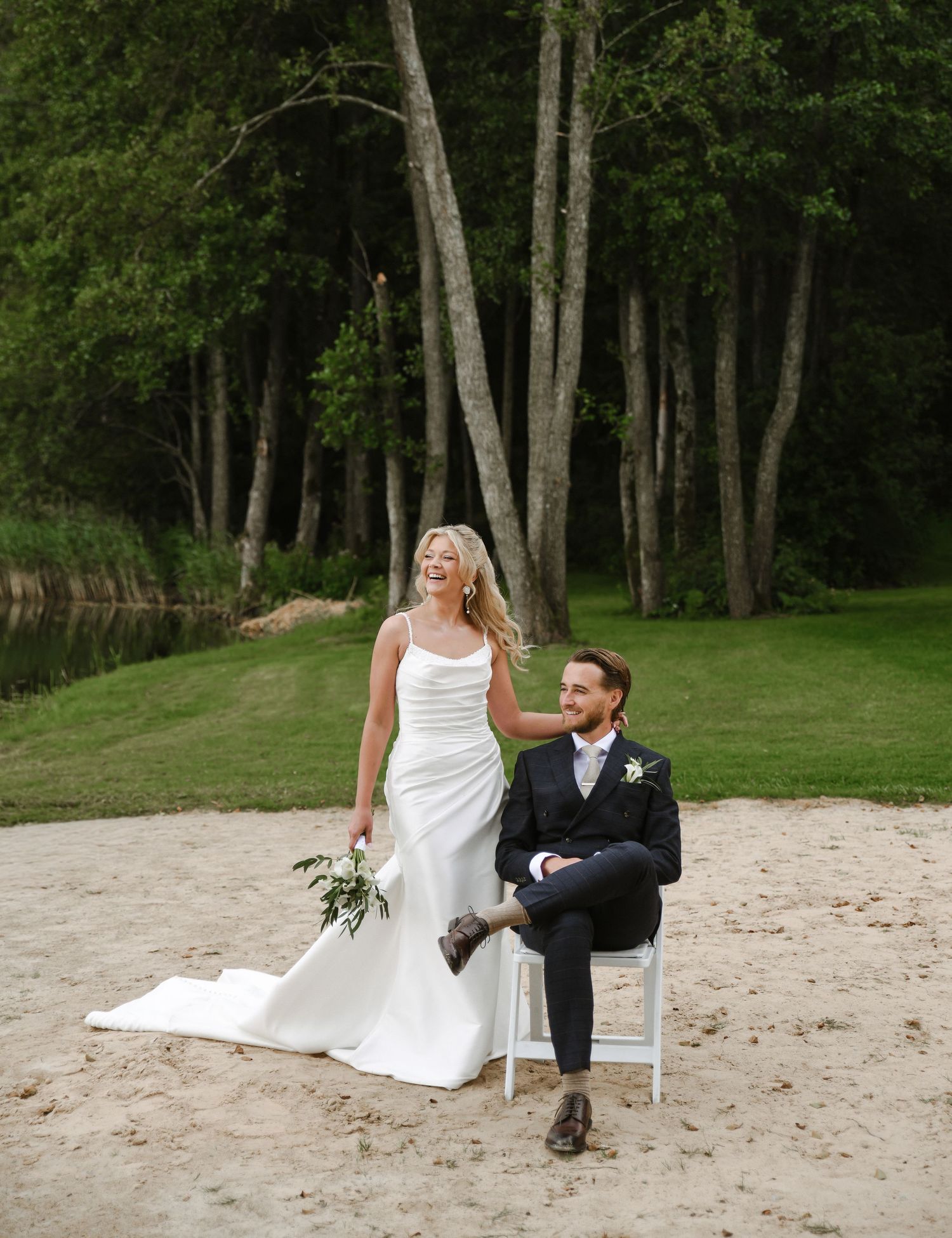 Wedding couple sits playfully together on a sandy beach with lush green trees in the background.