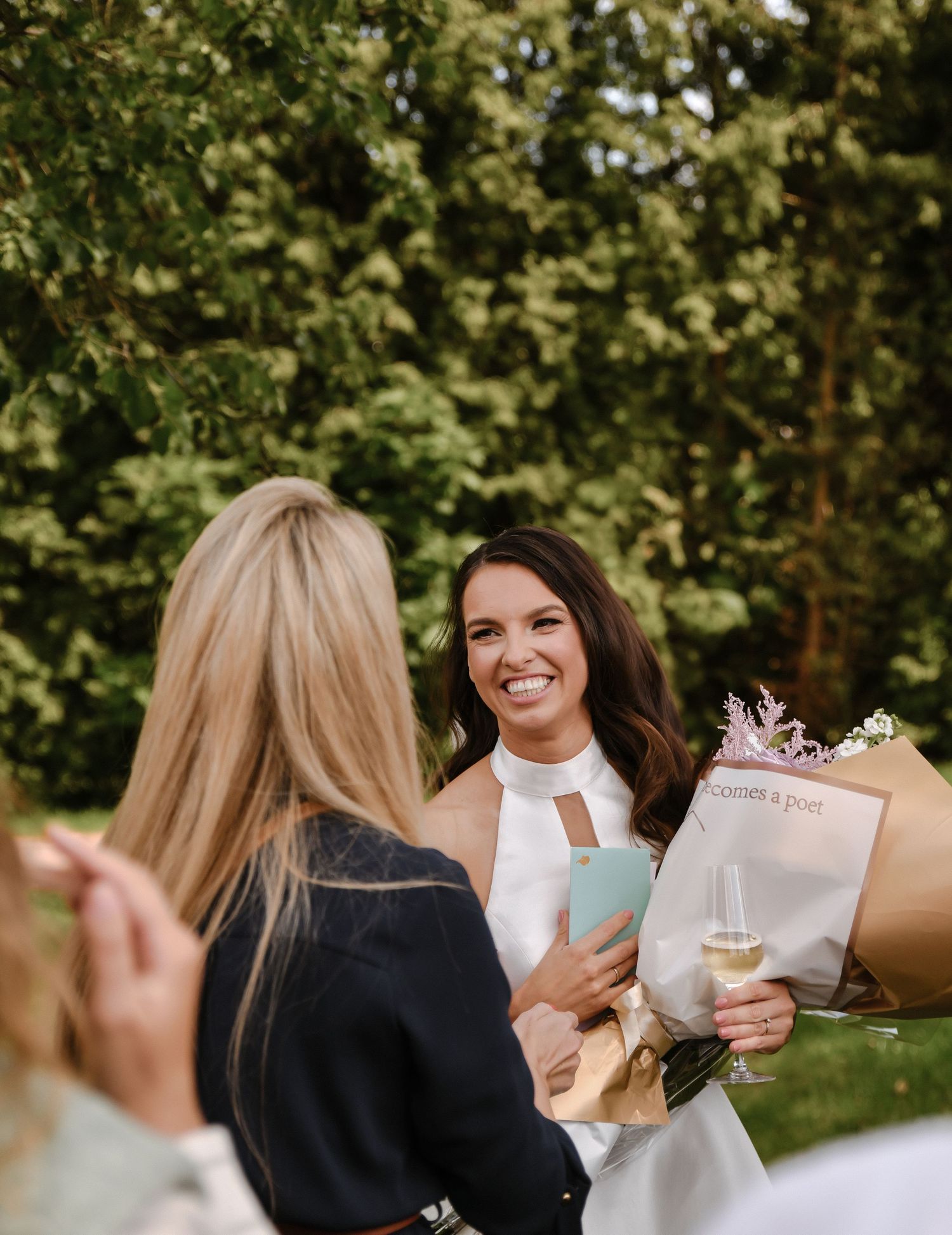 Two friends celebrate outdoors with champagne glasses and a floral bouquet wrapped in brown paper.