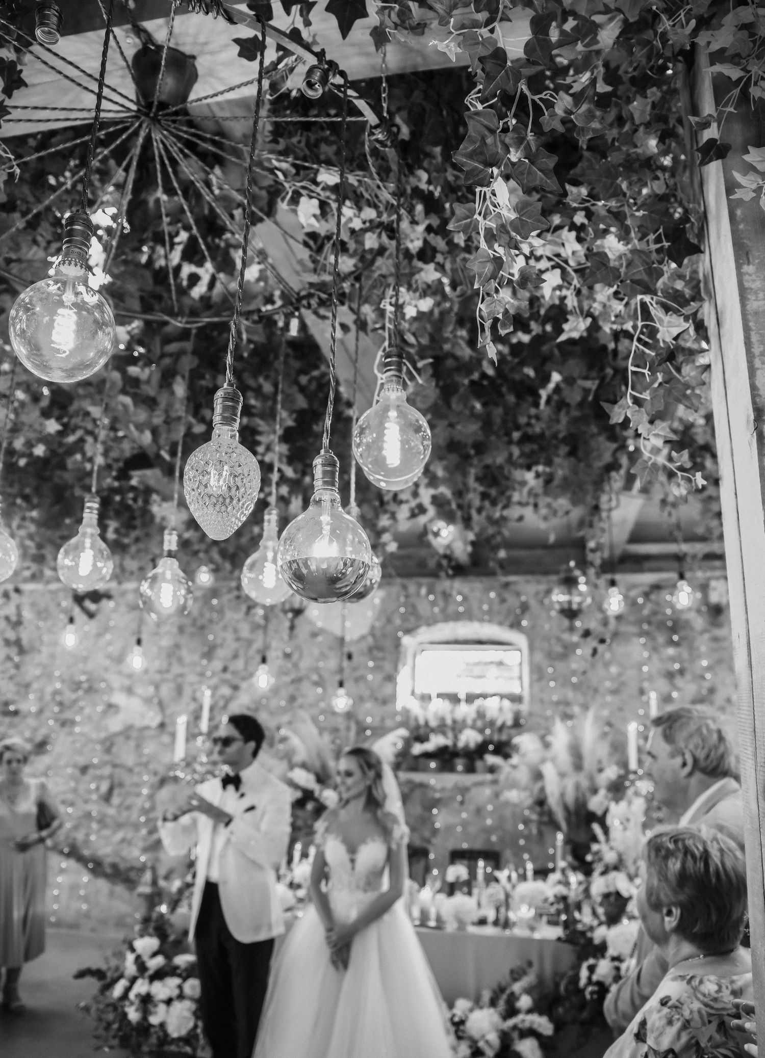 Black and white wedding photo showing glass bulb lights hanging from foliage over an elegant outdoor reception space.