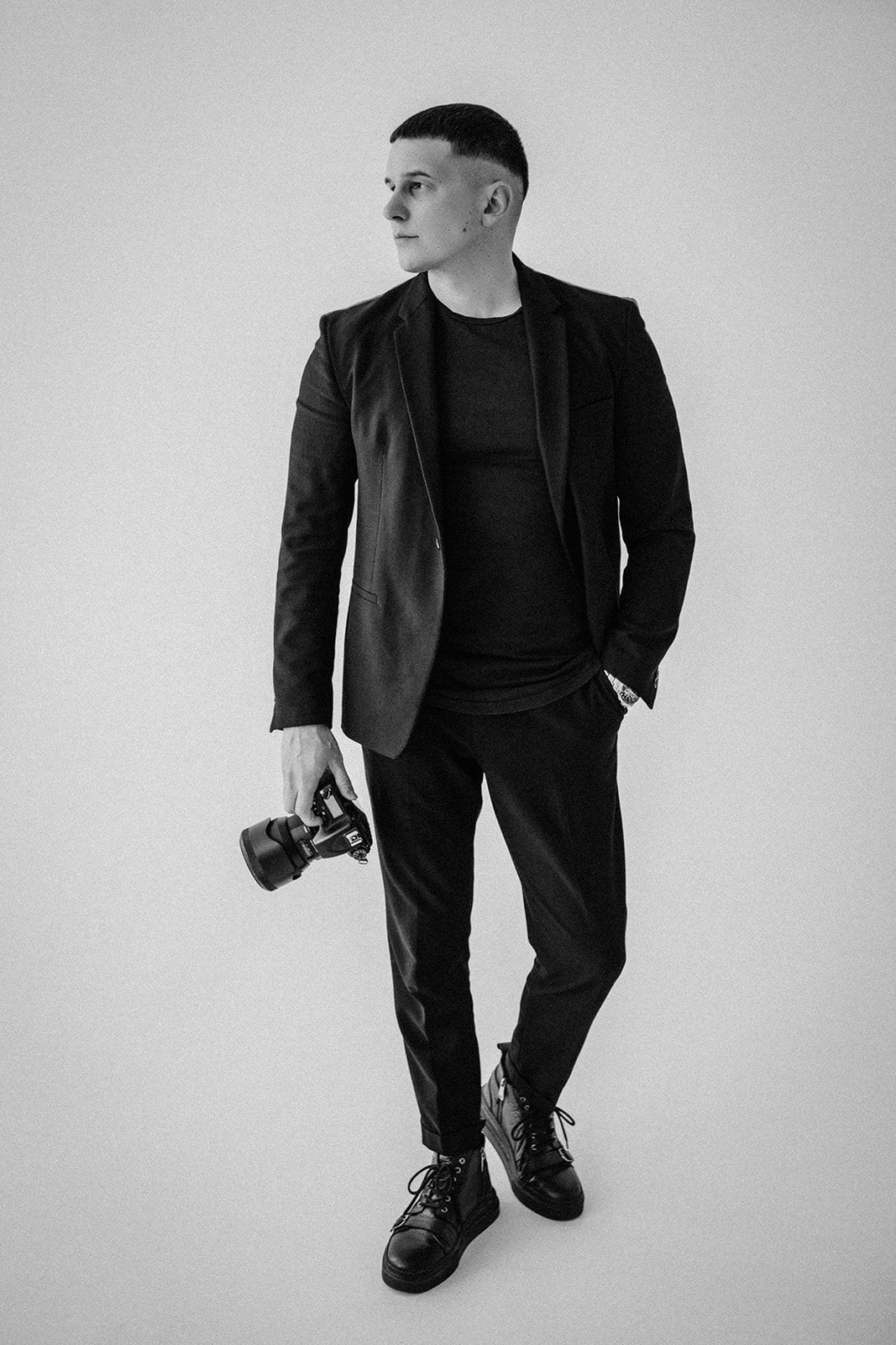 Professional photographer in black suit holds camera while posing against white backdrop in studio setting.