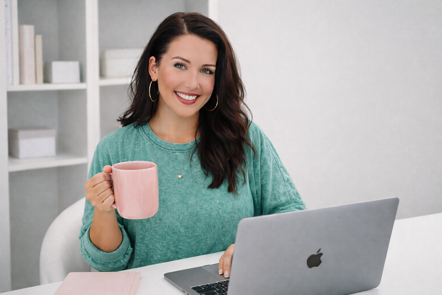 Person in teal sweater working on laptop while holding pink coffee cup in bright modern office setting.