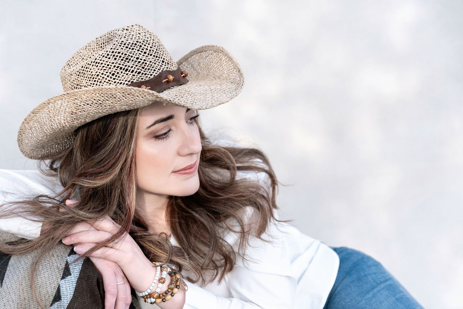 A carefree portrait of someone wearing a woven straw cowboy hat with flowing brown hair against a light background.