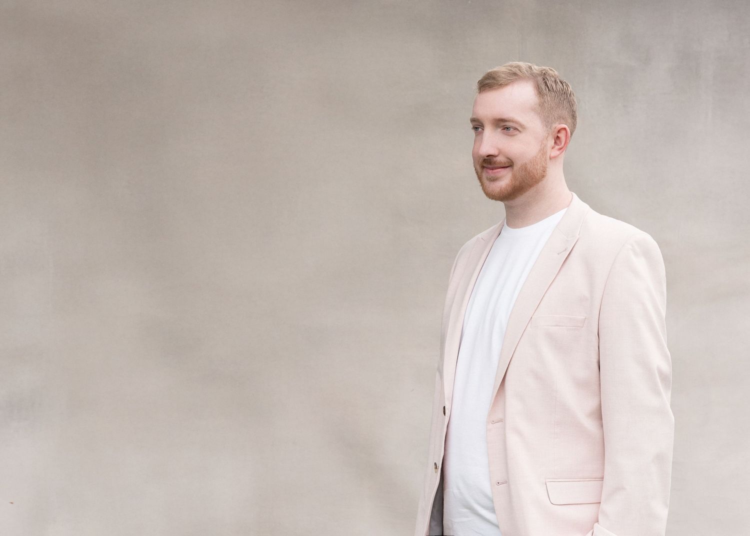 A person wearing a light beige blazer and white t-shirt stands against a neutral gray background with a subtle smile.