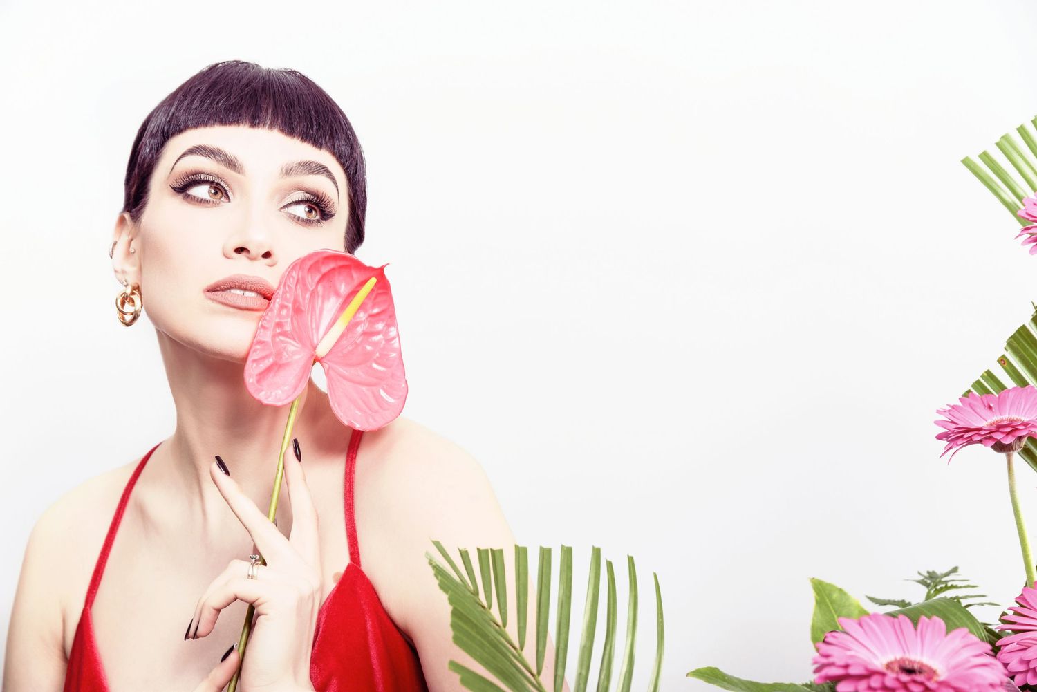 Woman in red dress poses with pink anthurium flower and tropical leaves against white background.