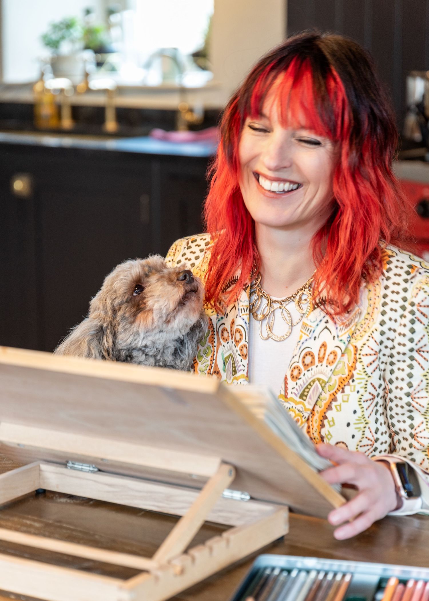 Someone with vibrant red hair smiles while reading a document with a small furry dog nearby in a bright kitchen setting.