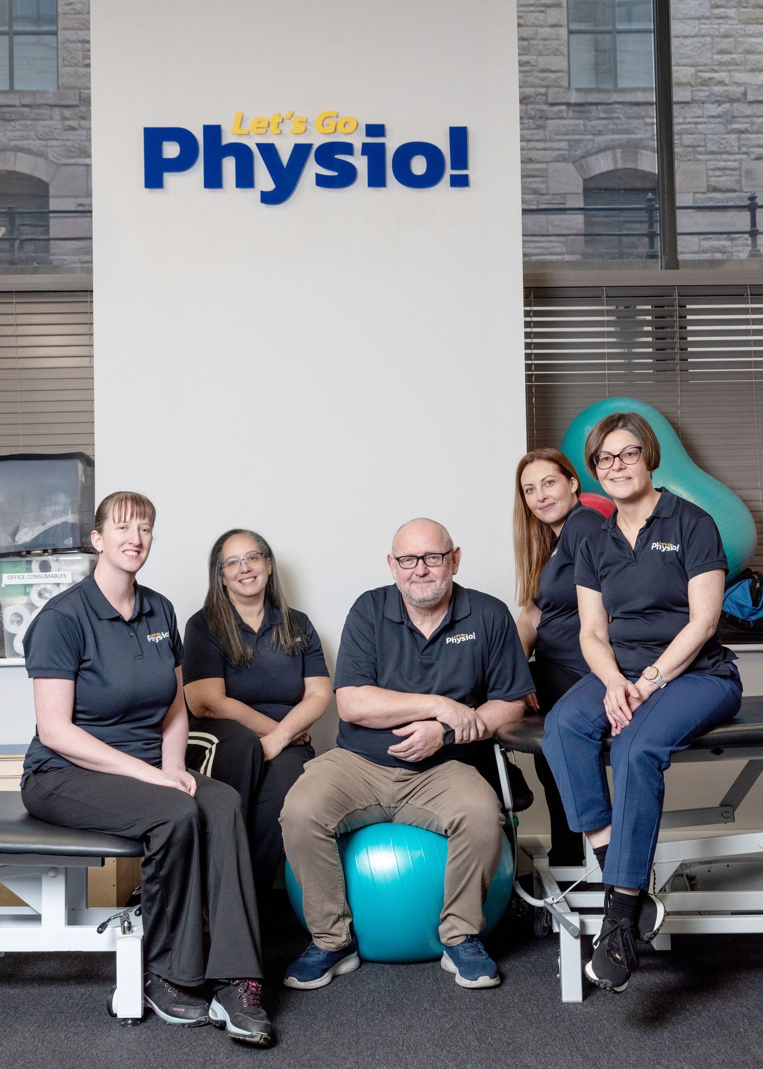 A group of physiotherapy staff members sit together on treatment tables with exercise equipment in a medical office.