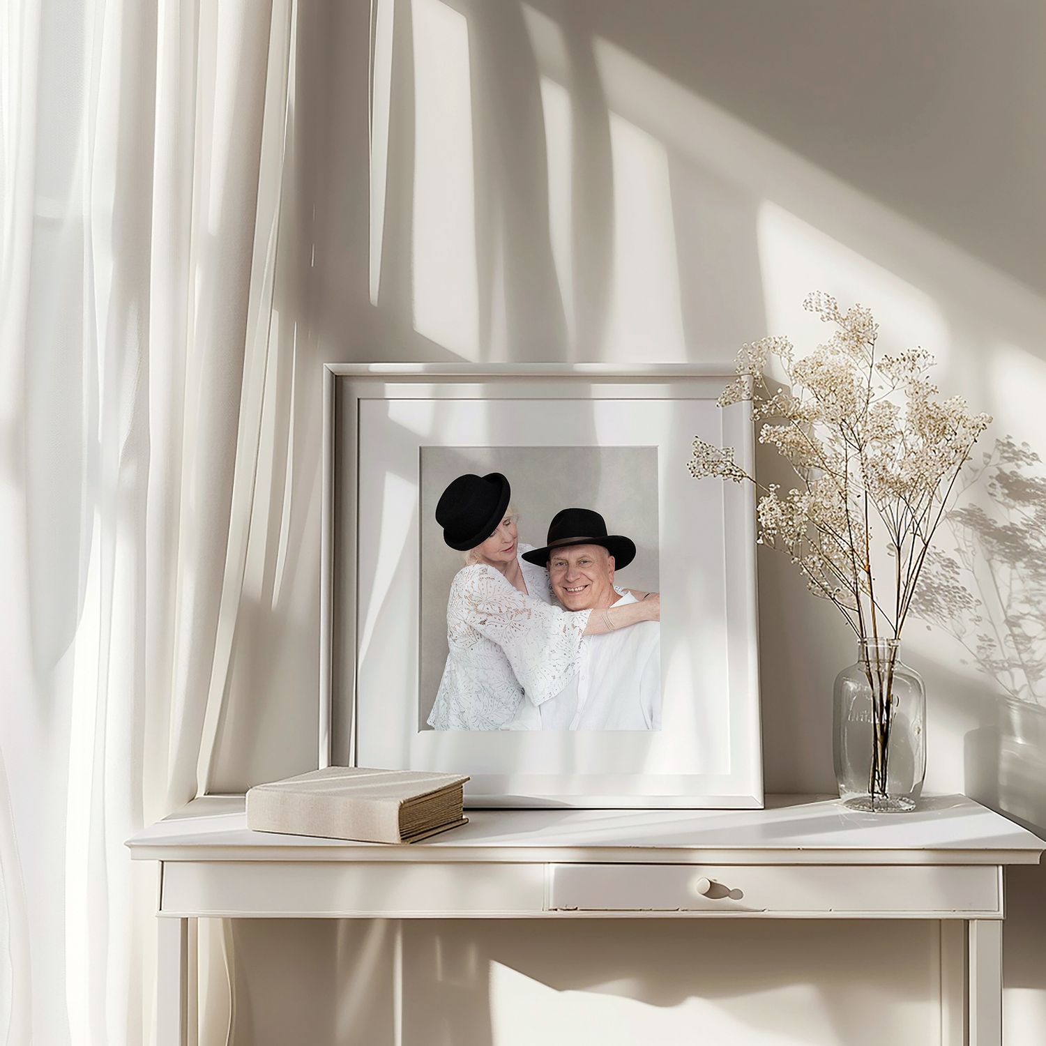 Framed black and white photograph on white desk with dried flowers and sunlit curtains creating soft shadows.