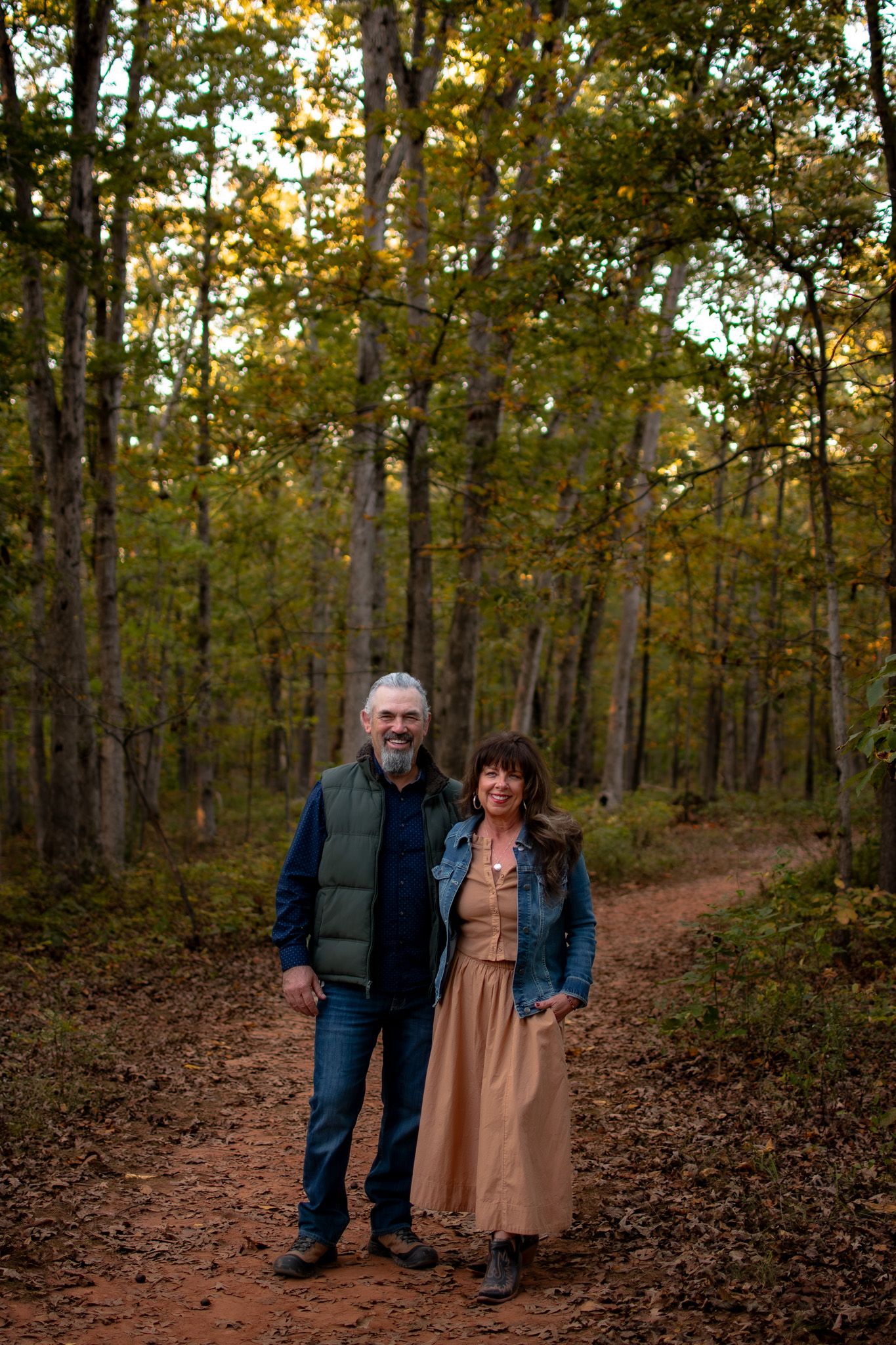 Two people stand together on a leaf-covered forest path during autumn with golden sunlight filtering through trees.