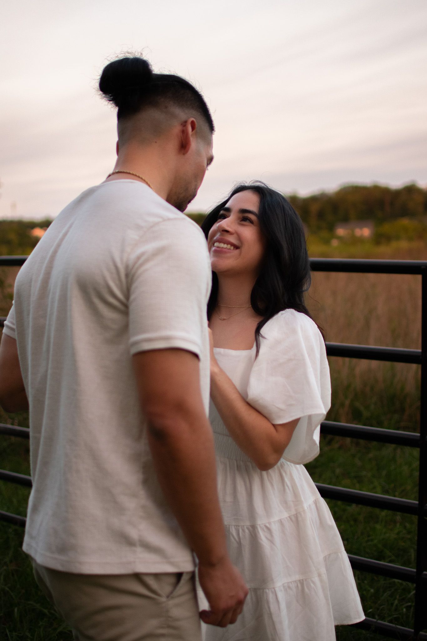 A romantic couple in white outfits share tender moments while standing by a wooden fence at sunset.
