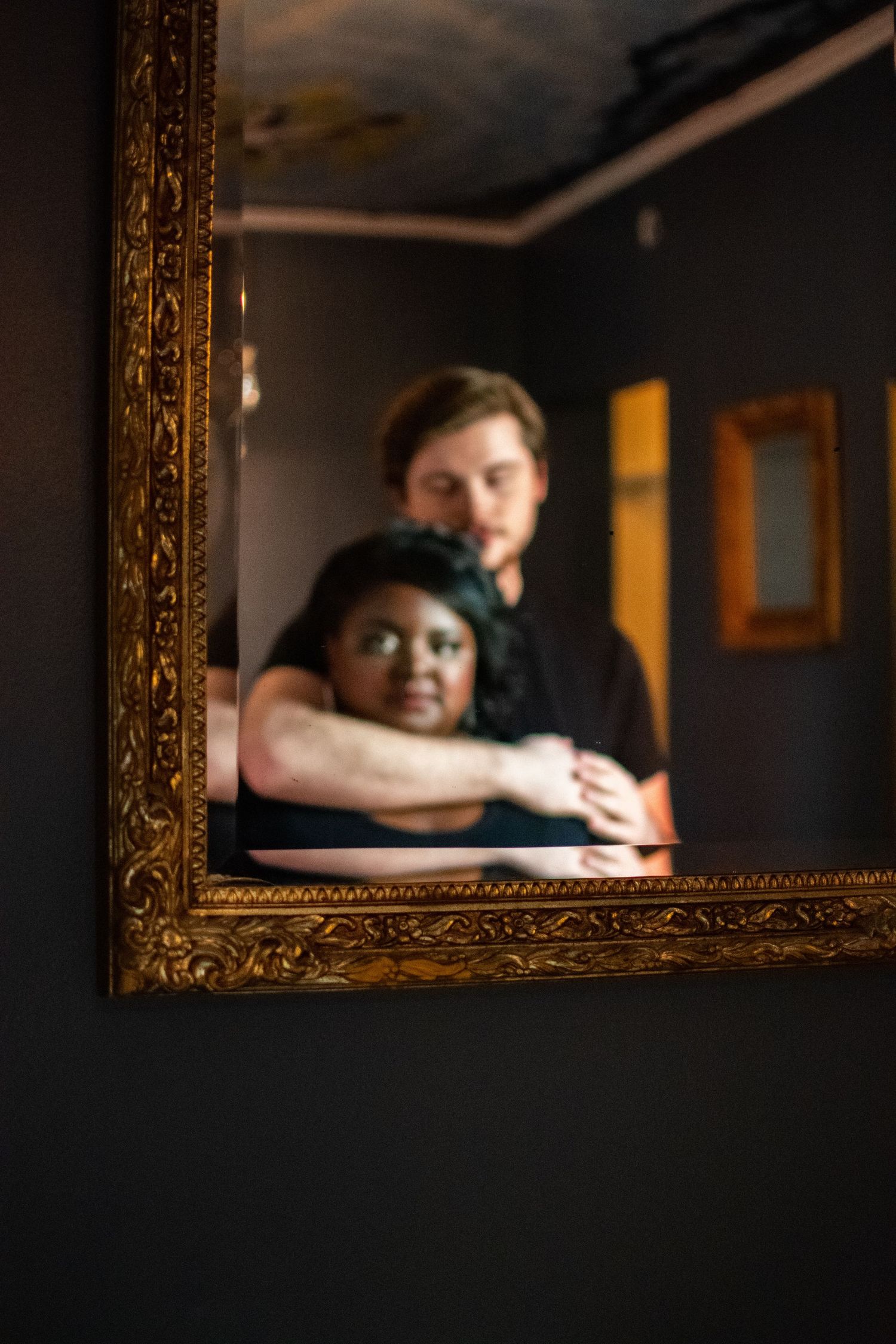 A creative portrait composition showing two people reflected in an ornate gold frame mirror against a dark background.