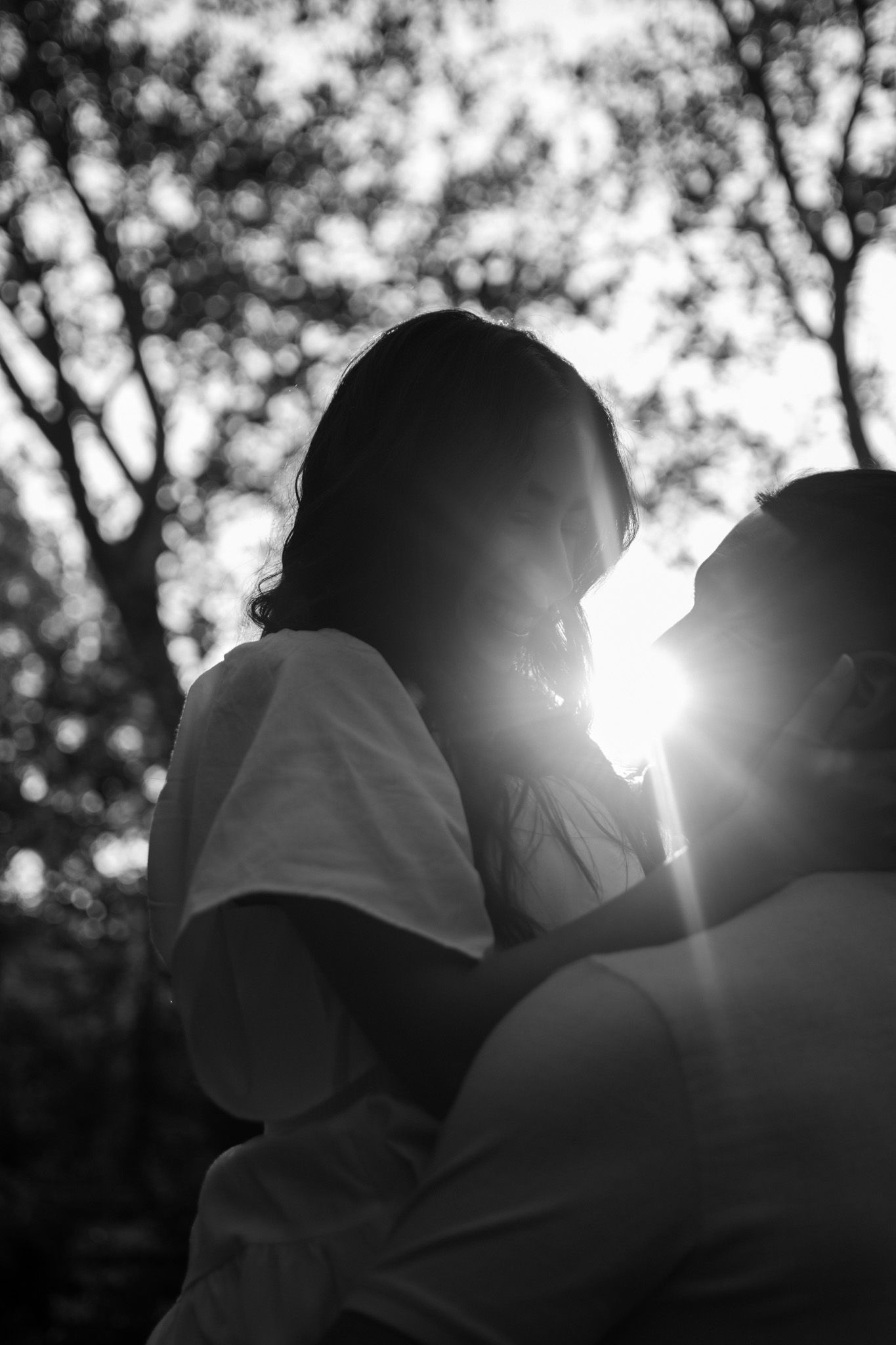 Silhouetted figures stand against bright sunlight filtering through trees in a black and white photograph.