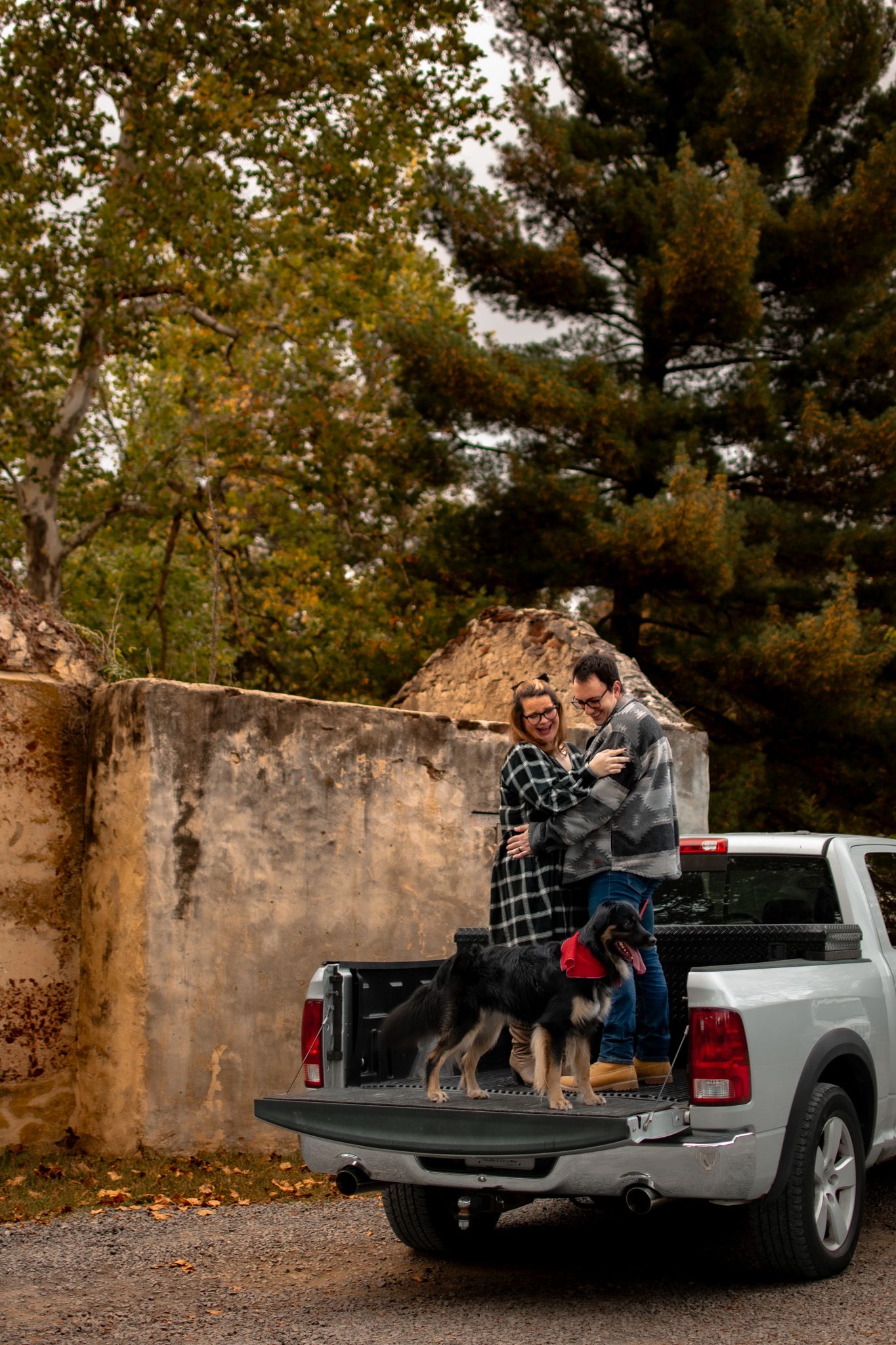 A couple sits together in the back of a silver pickup truck during an autumn photoshoot against a stone wall.