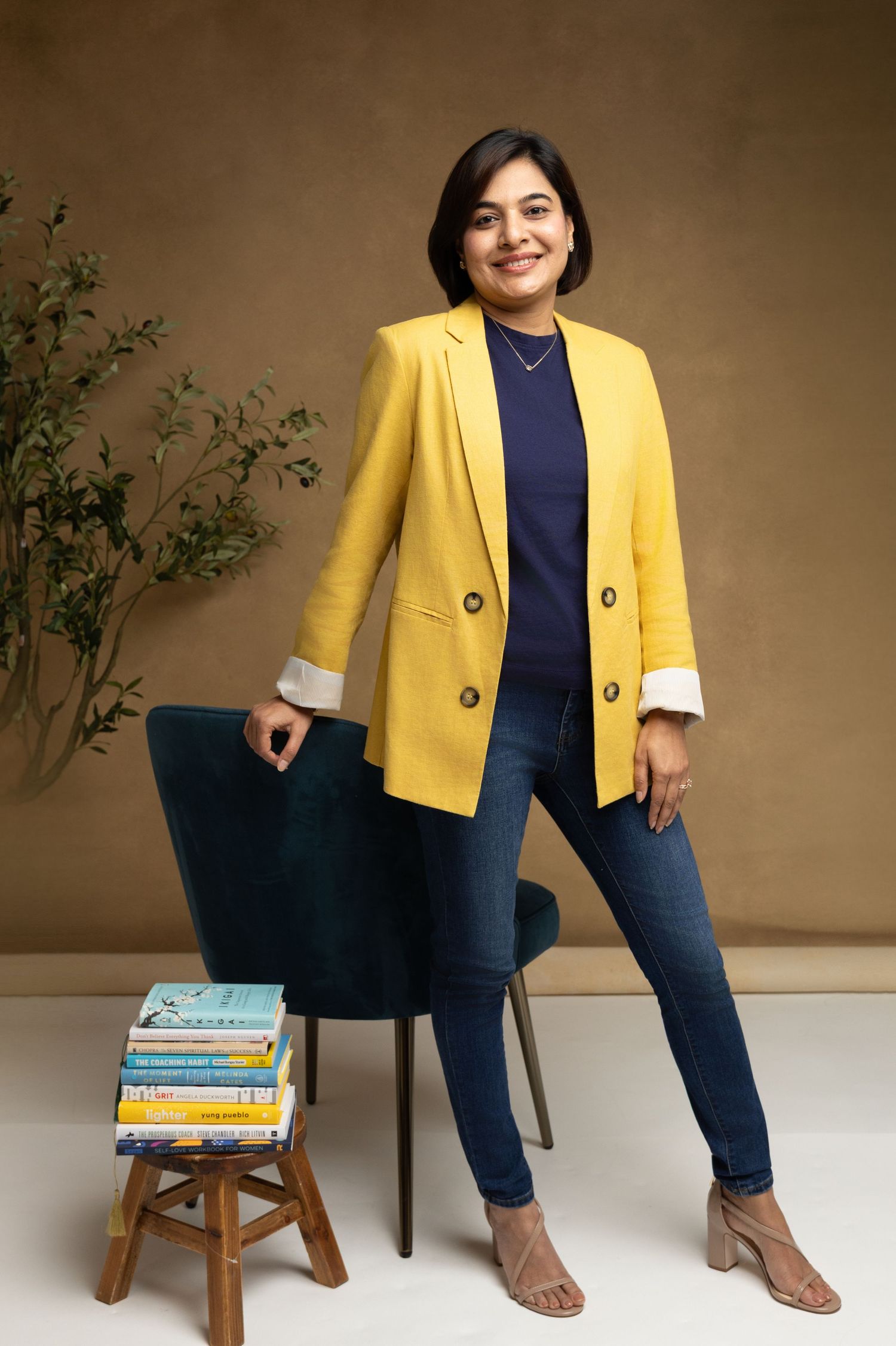 Woman in yellow blazer and jeans poses confidently beside a stack of books and teal chair in a studio setting.
