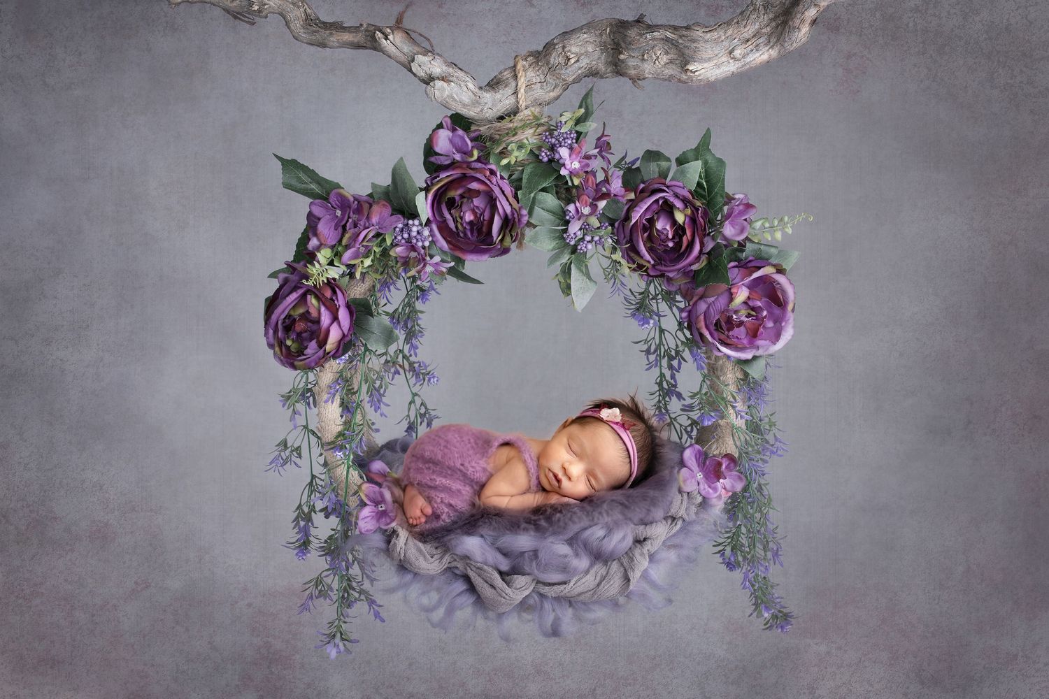 Newborn baby sleeping in a purple floral wreath swing against a gray backdrop in a professional photo.