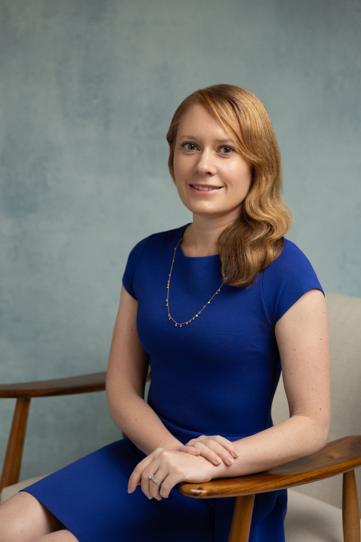 Professional woman in blue dress seated in wooden chair, smiling confidently against gray studio background.