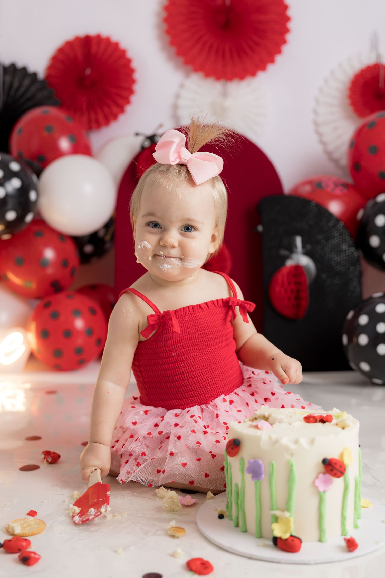 A festive first birthday cake smash photo with red and white decorations, balloons, and paper fans in the background.
