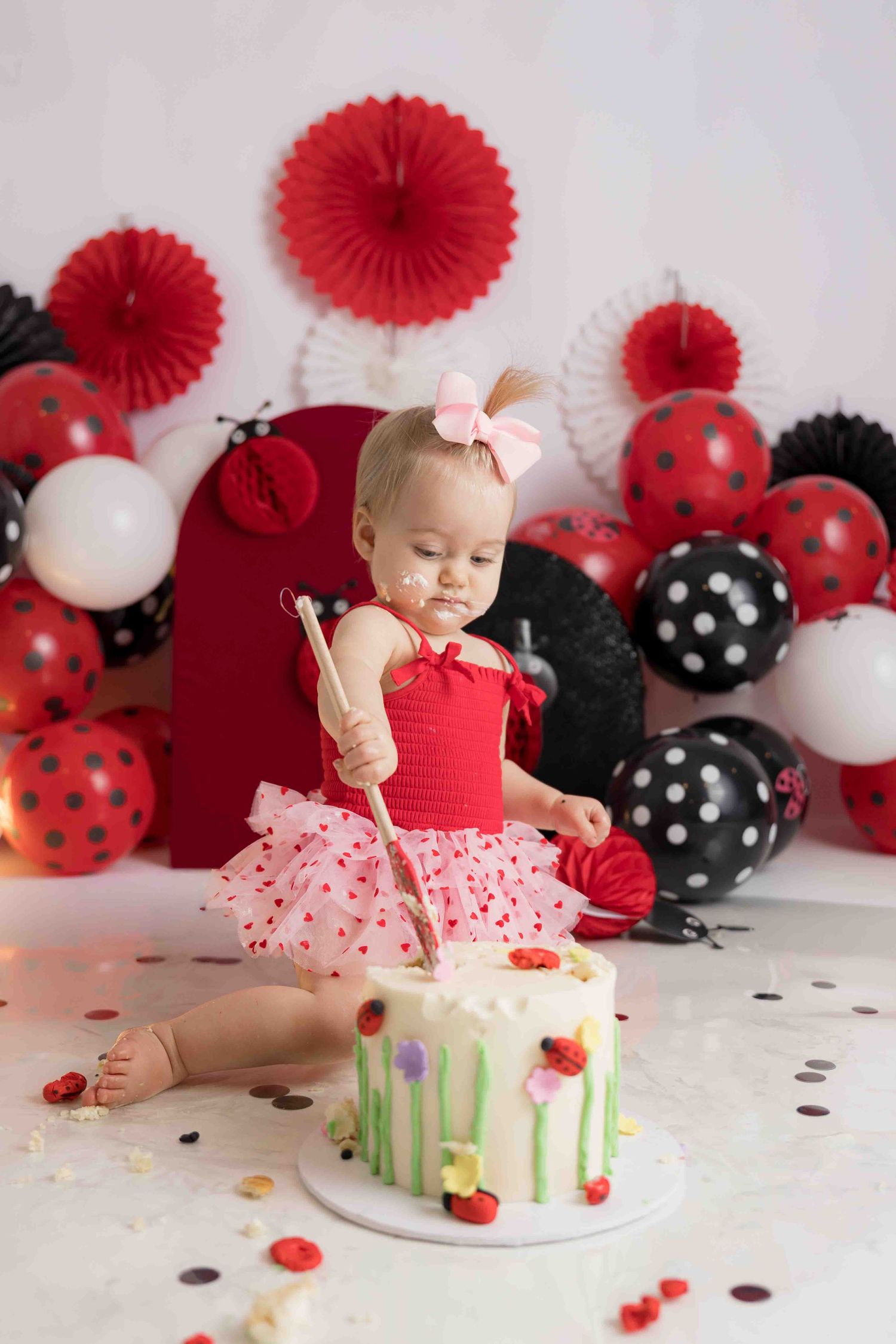 Cake smash photo session with red and black polka dot decorations, paper fans, and balloons against white backdrop.