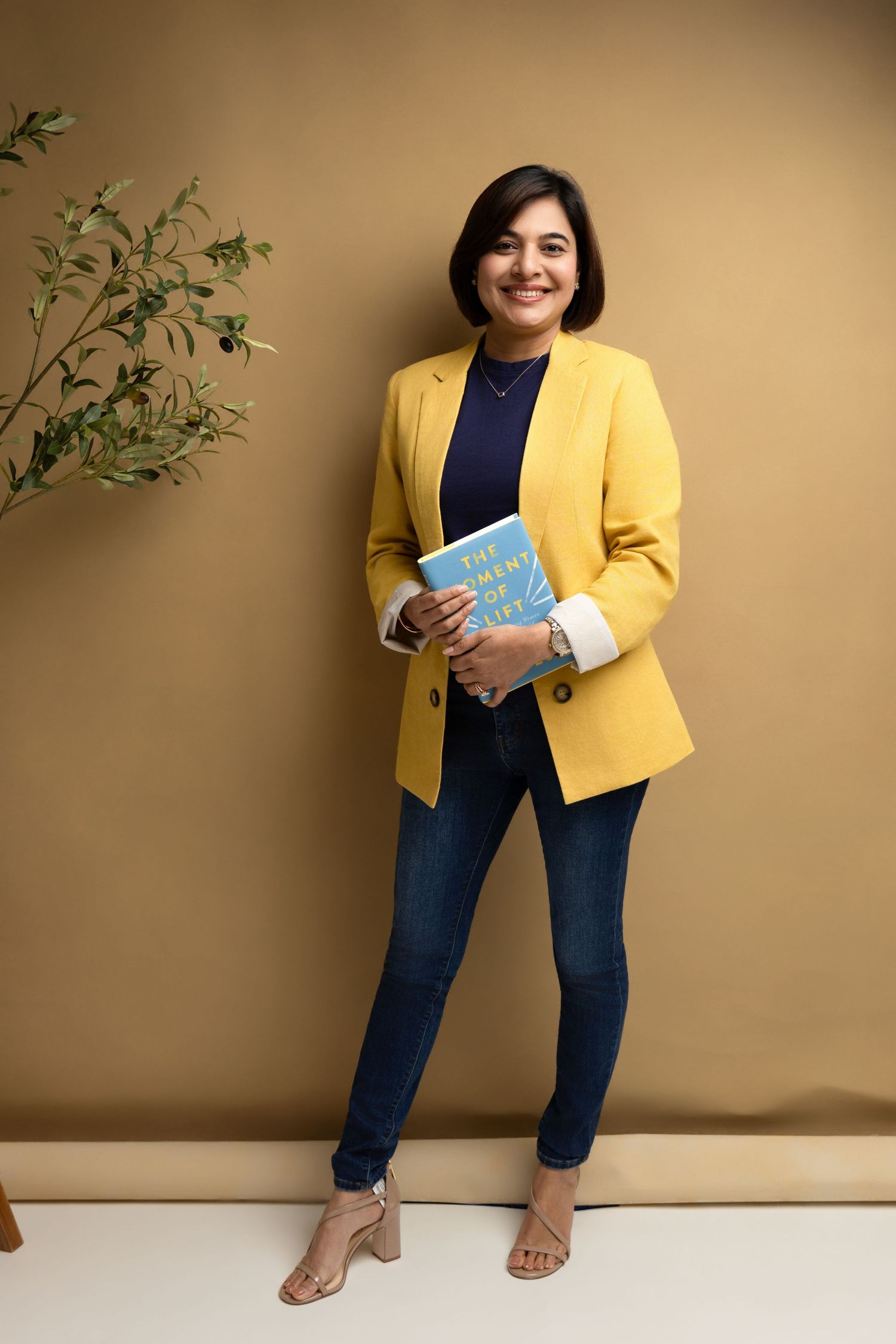 Woman in yellow blazer and jeans smiling while holding a blue book, standing against a tan wall.