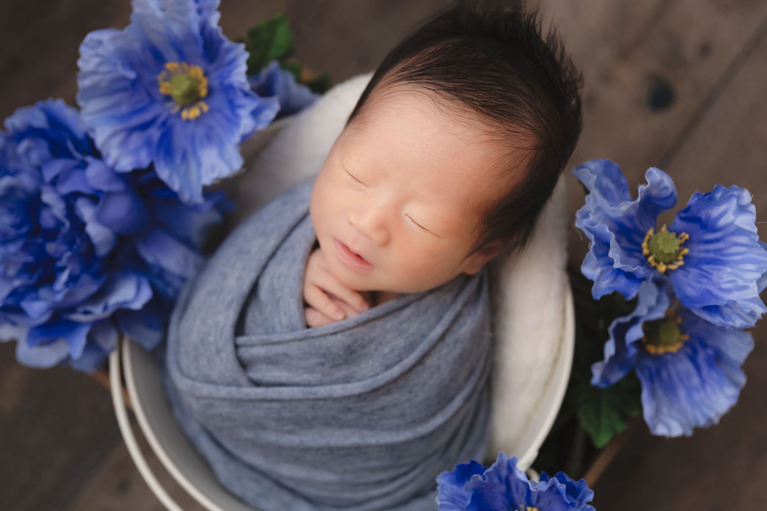 Newborn wrapped in grey swaddle surrounded by blue poppies on wooden surface.