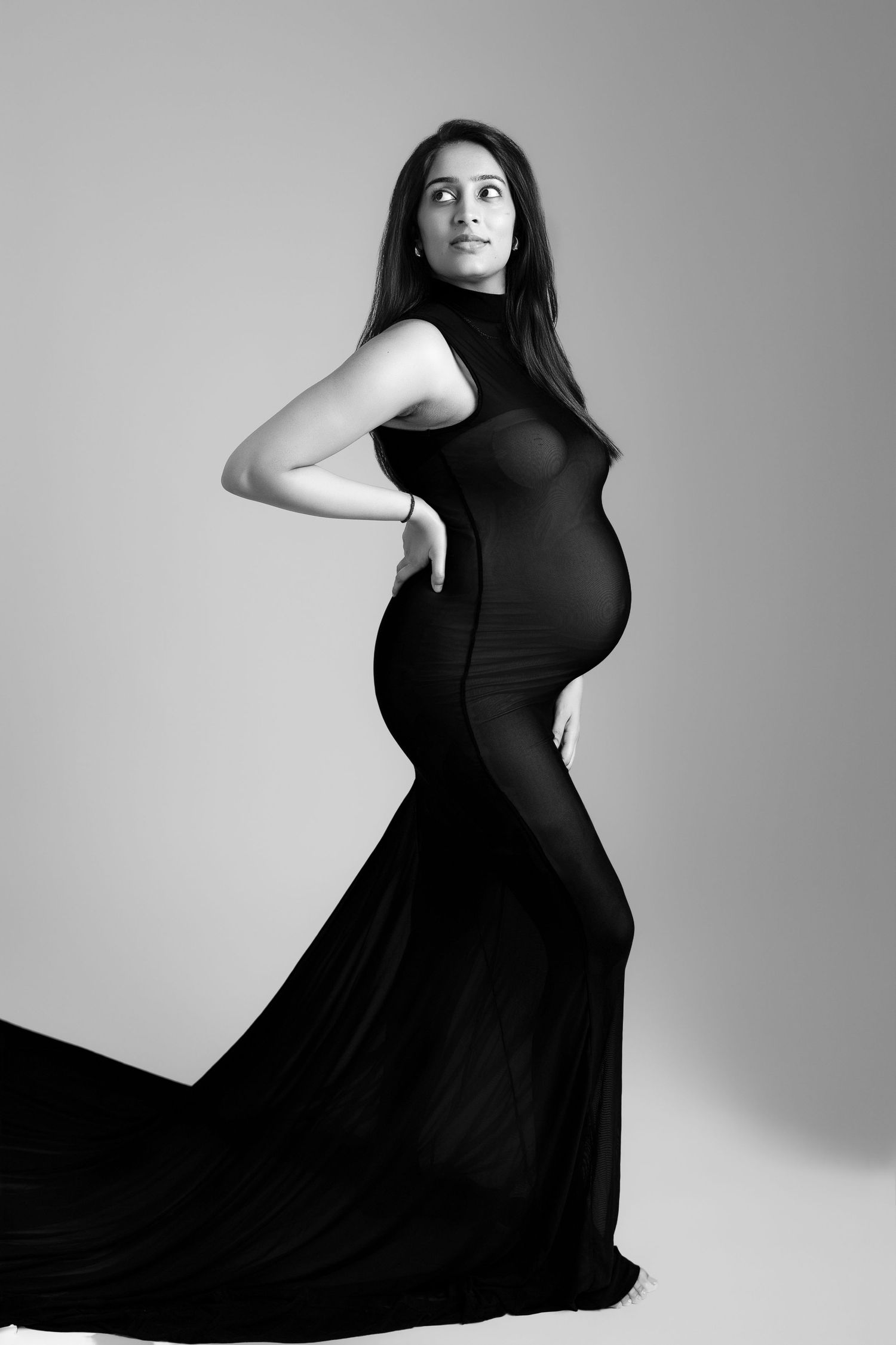 Black and white photo of a pregnant woman in an elegant black gown, posing gracefully in a studio setting.