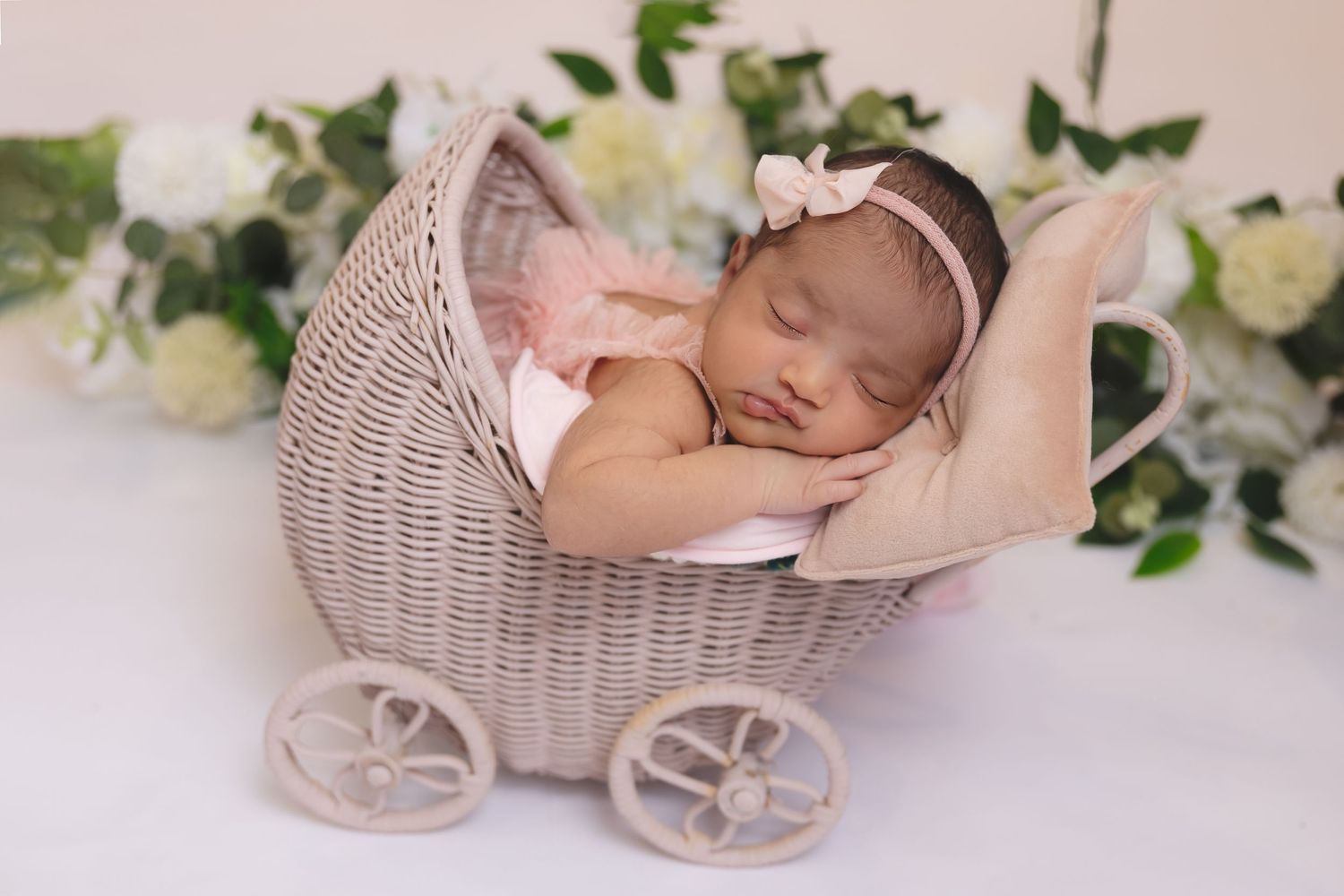Sleeping newborn in miniature wicker carriage with floral decorations.