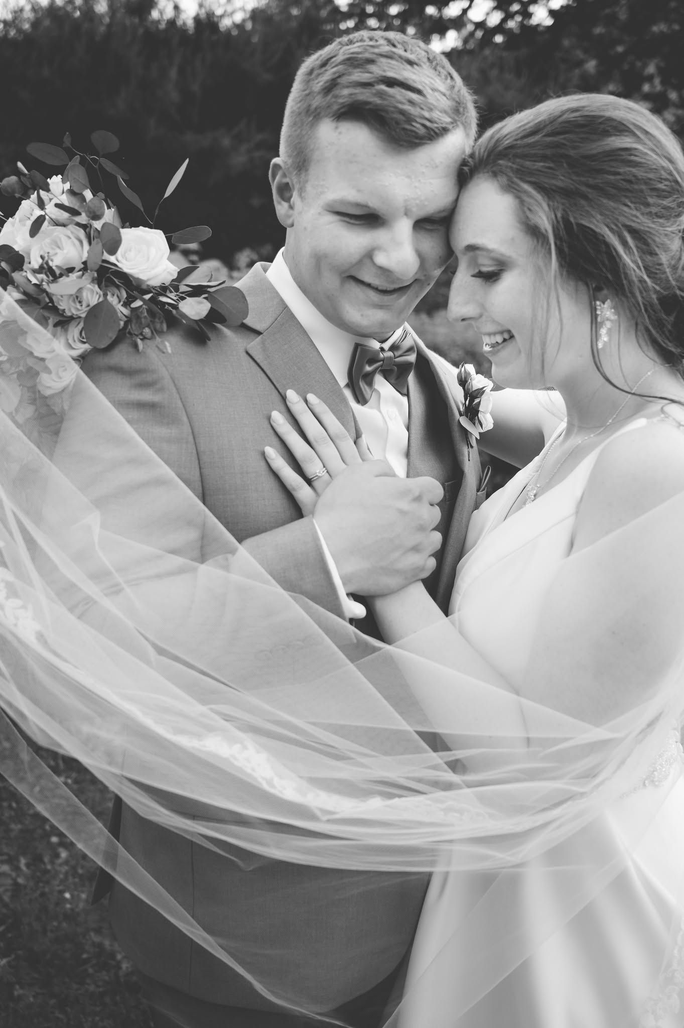 Black and white wedding photo of smiling bride and groom embracing, veil flowing, bride holding bouquet.