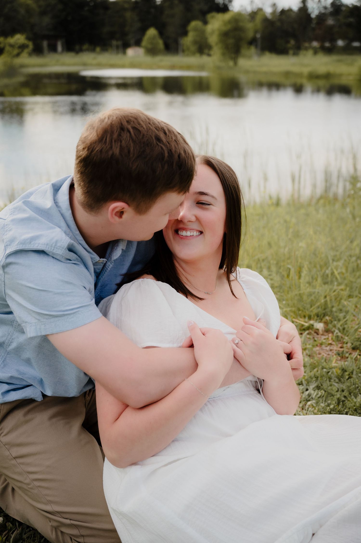 Couple sharing a romantic moment by a serene lake, woman in white dress smiling joyfully as man embraces her.